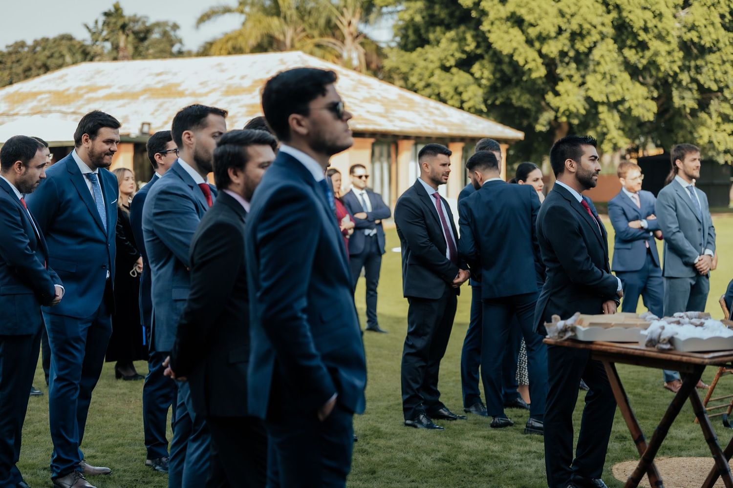 Group of men in navy suits standing on green lawn at outdoor wedding venue.