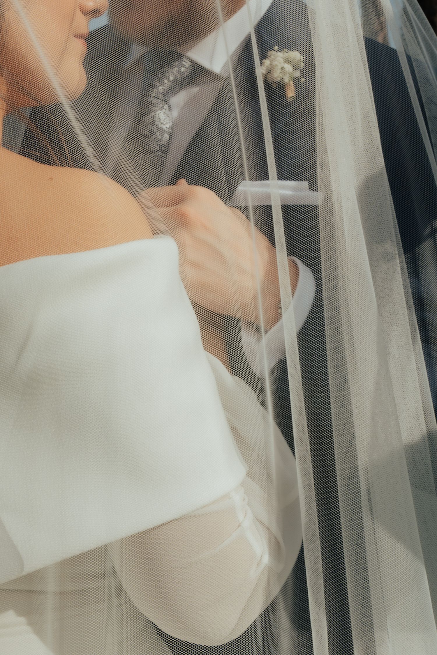 Close-up of bride in off-shoulder white gown and groom in suit embracing under a sheer veil.