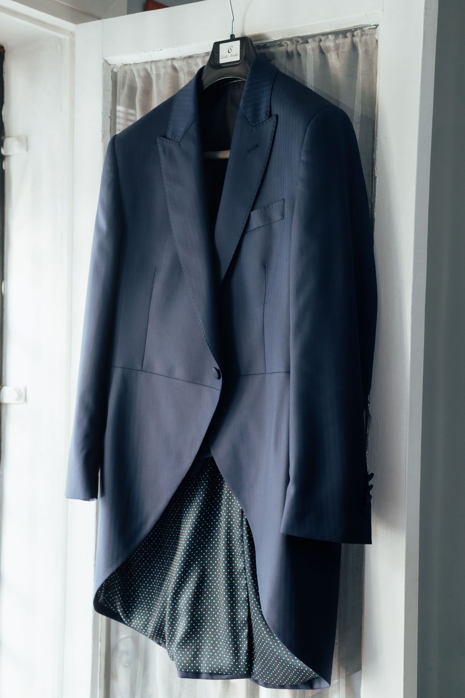 Navy blue tailcoat suit jacket hanging on hanger in front of window.