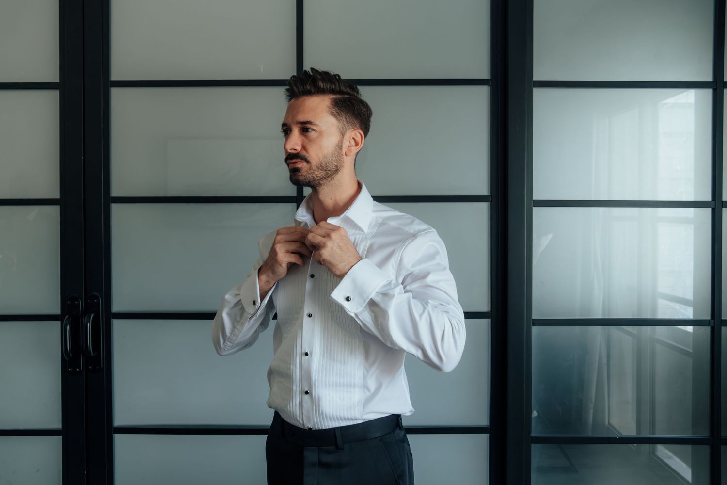 A well-dressed man in a white formal shirt adjusts his collar buttons while getting ready indoors.