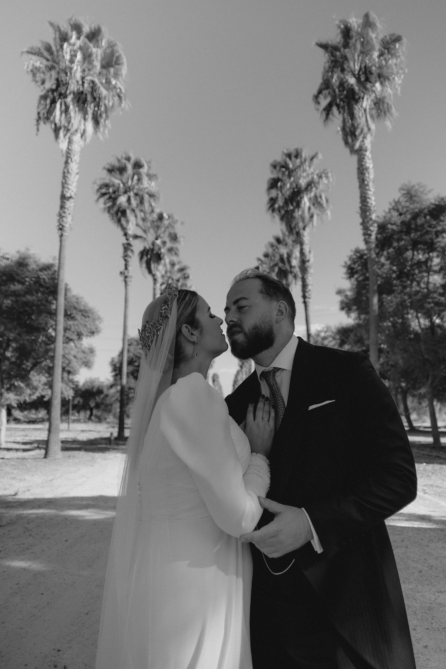 Black and white photo of bride and groom embracing romantically among tall palm trees on their wedding day.