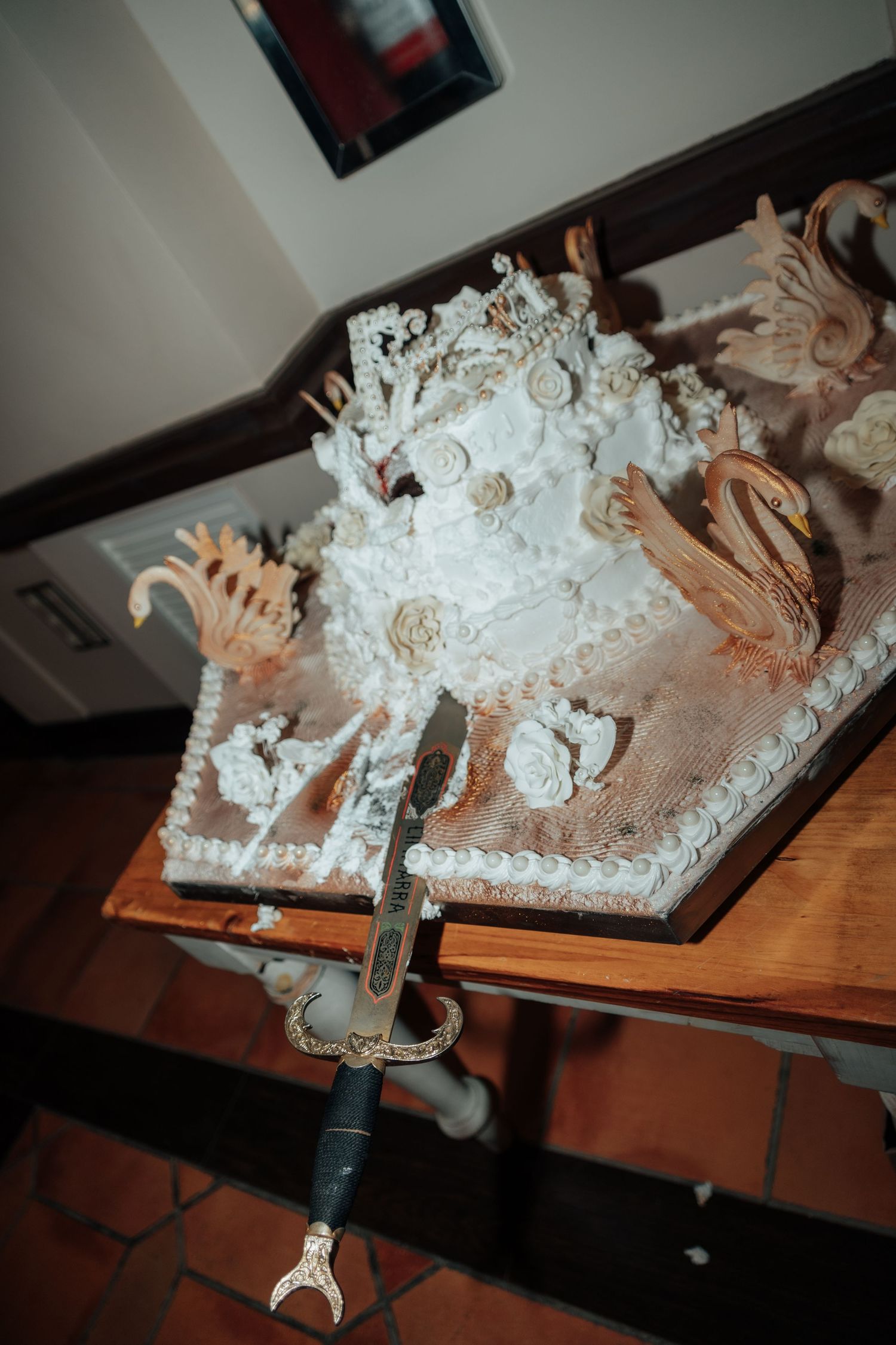 Ornate white wedding cake decorated with swans and roses with ceremonial sword on table.
