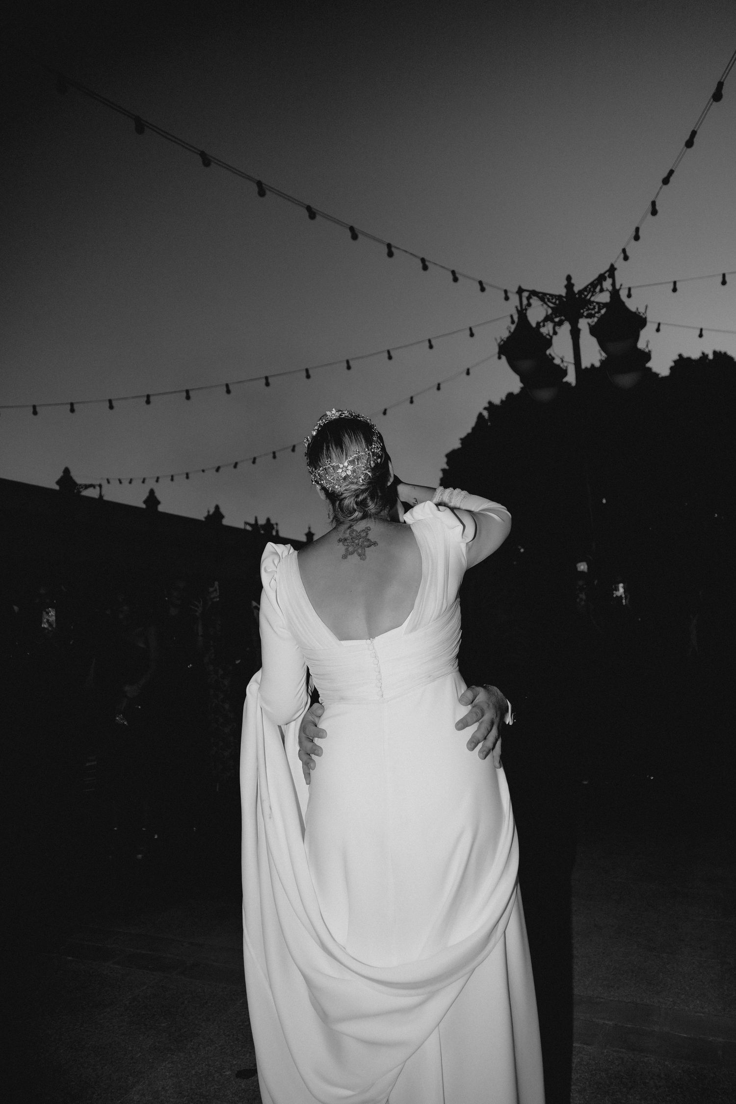 Bride in elegant white gown with back tattoo dancing under string lights at outdoor wedding reception.