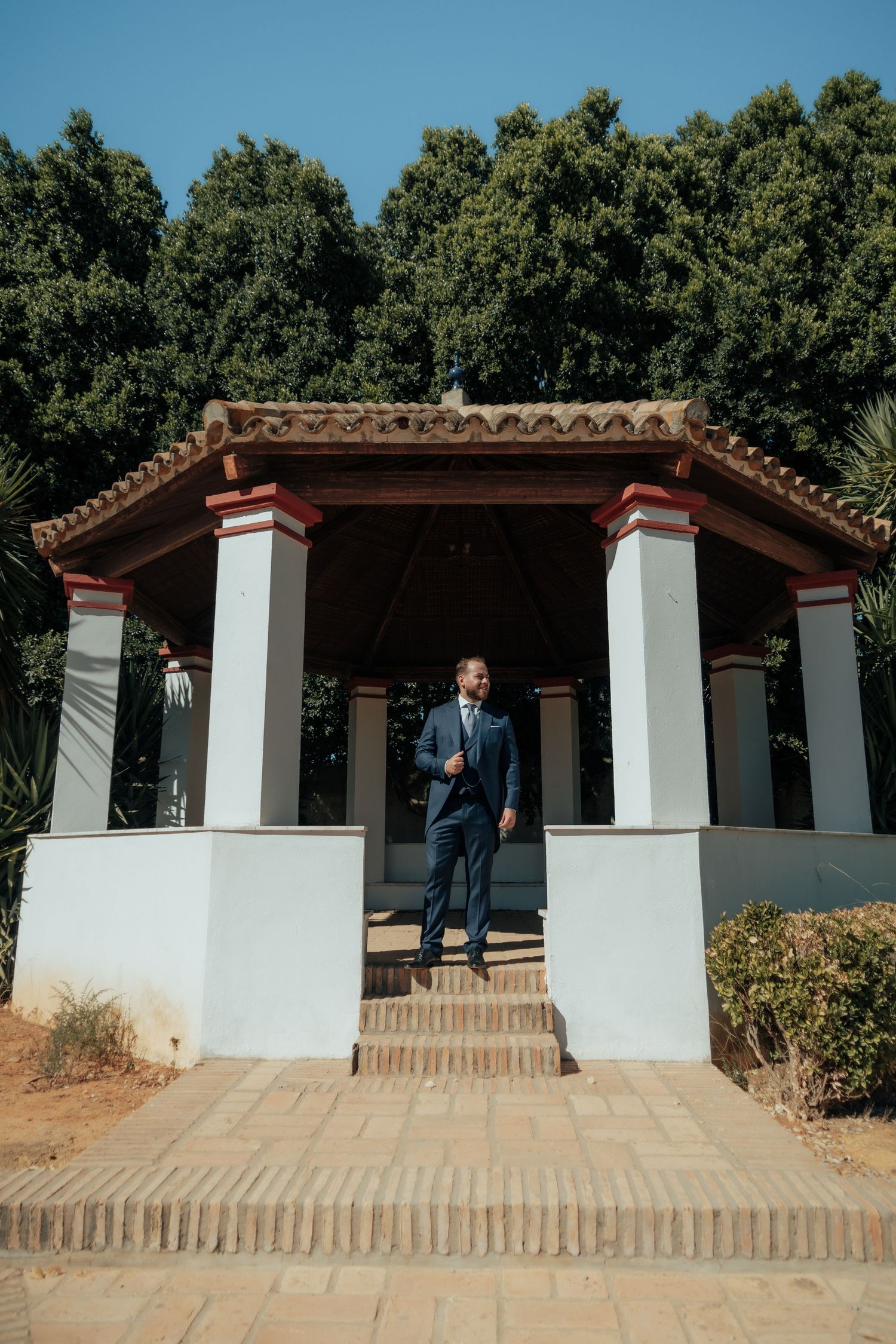 Dapper groom in navy three-piece suit stands confidently under a white Spanish-style pavilion outdoors.