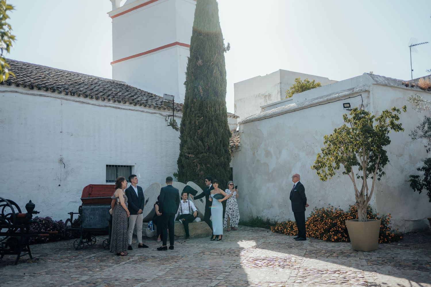 Wedding guests mingling in a whitewashed Spanish courtyard with a cypress tree and church tower.