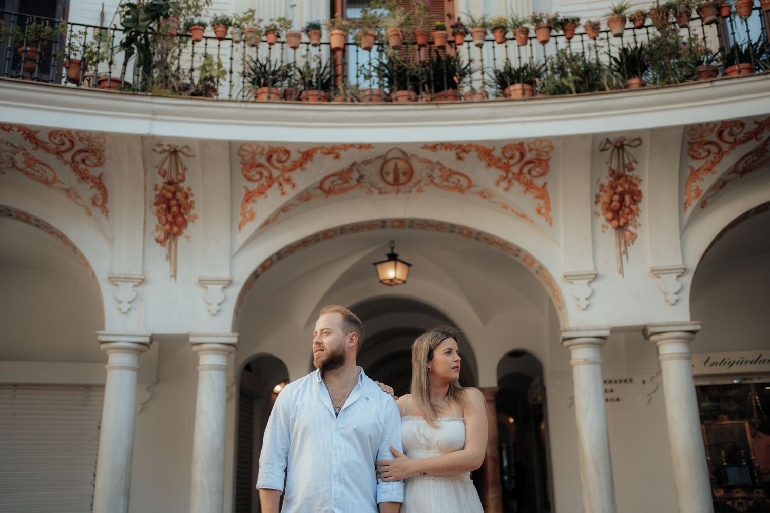 Couple embracing under ornate white arches with decorative balcony and potted plants in Seville Spain.