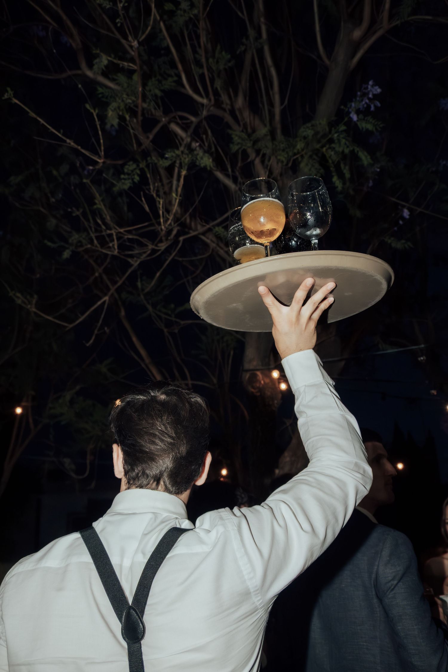 Waiter in white shirt and suspenders balancing a tray of drinks overhead at outdoor night event.