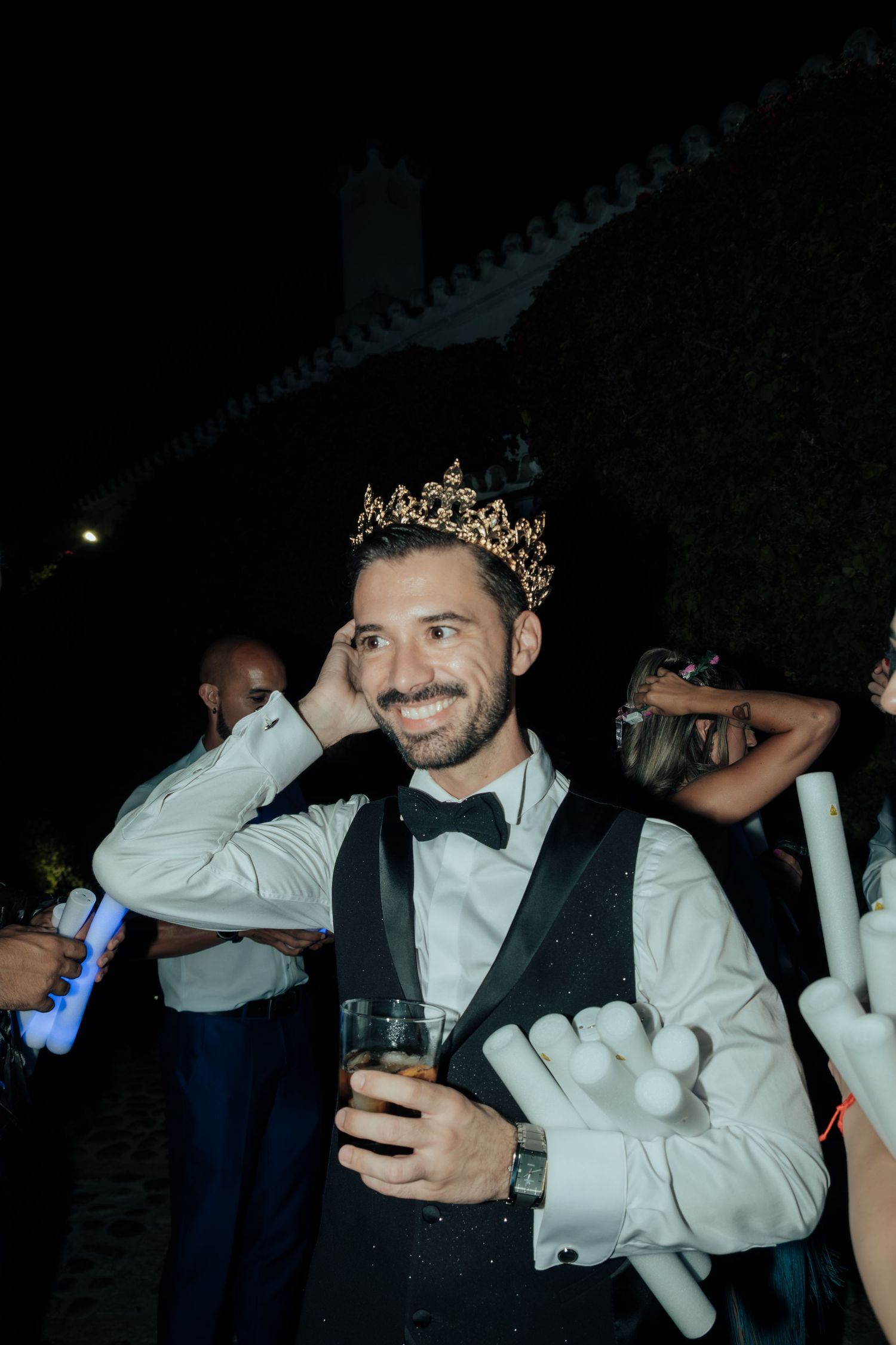 Smiling groom wearing gold crown and bow tie holding drink at outdoor wedding celebration at night.