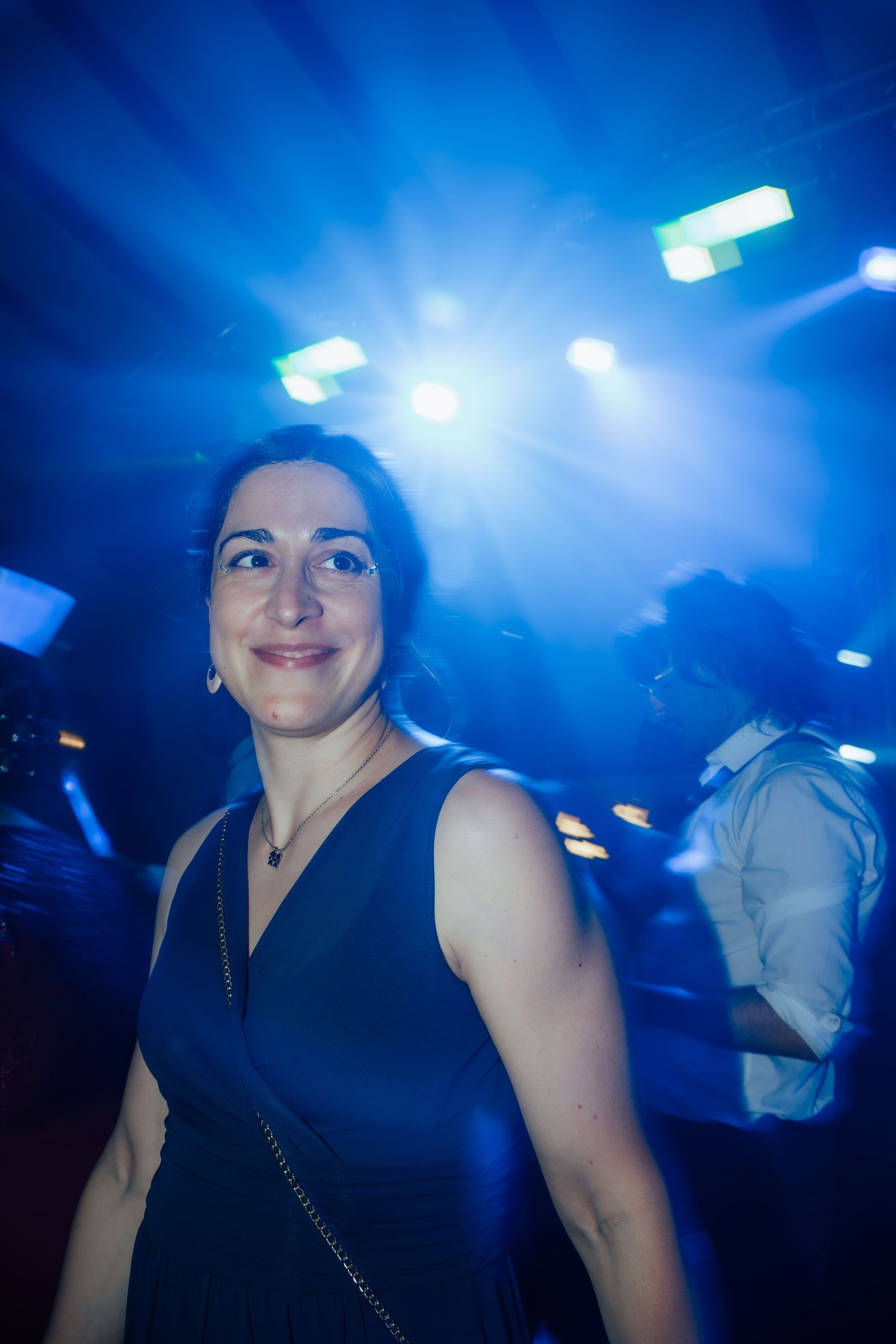 A smiling woman in a black dress poses on a dance floor illuminated by dramatic blue stage lighting.