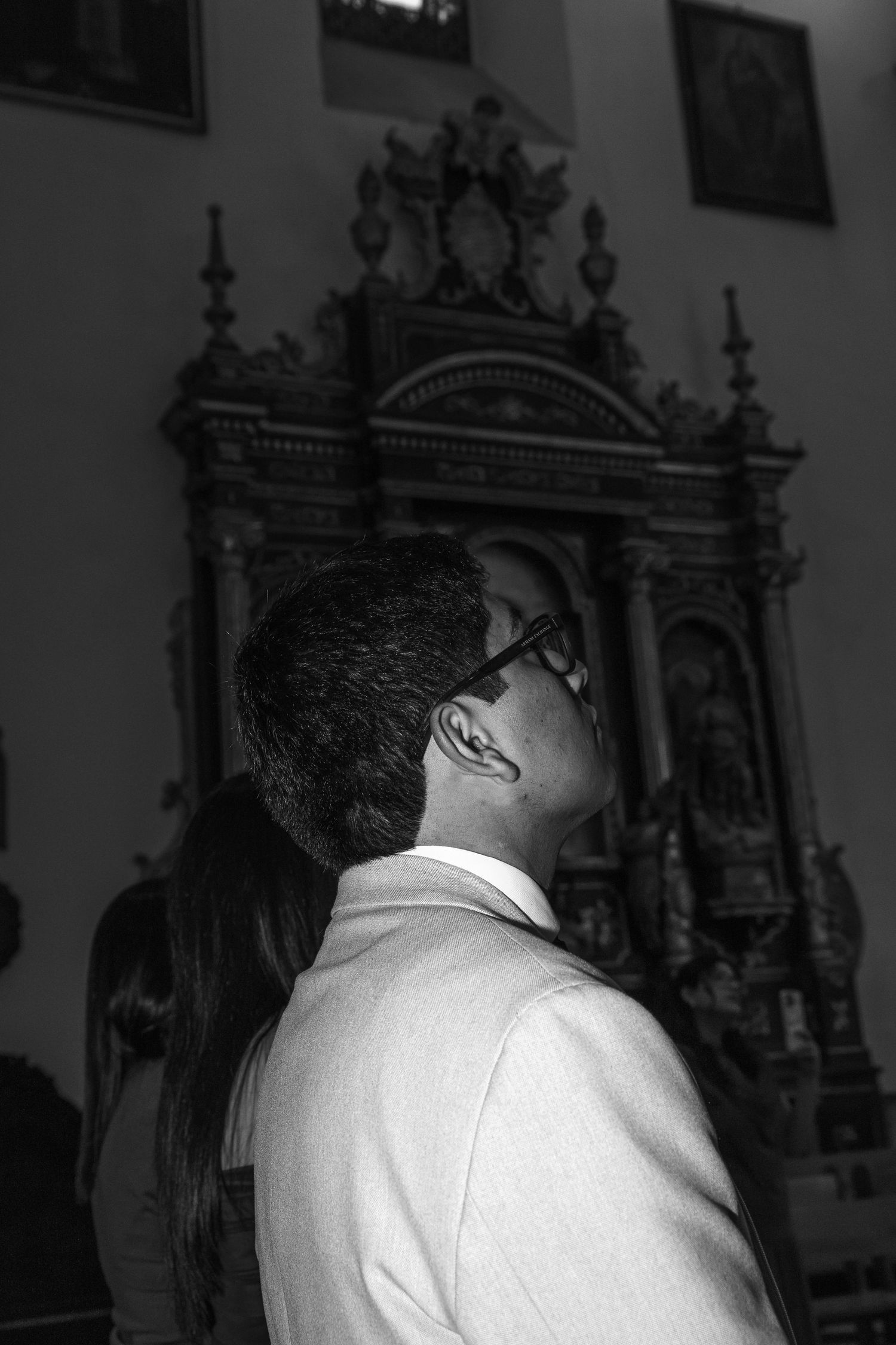 Black and white portrait of groom in suit gazing up at ornate church altar during ceremony.
