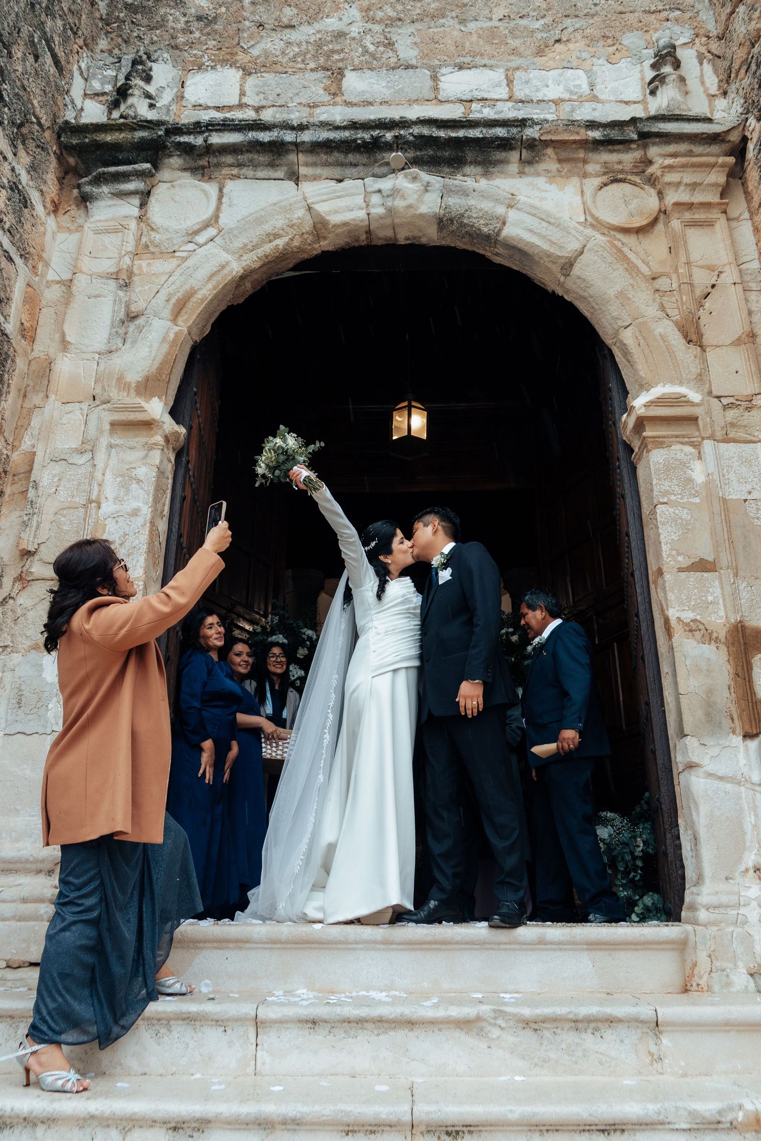 Newlyweds share a kiss on stone church steps as guest photographs them after the ceremony.