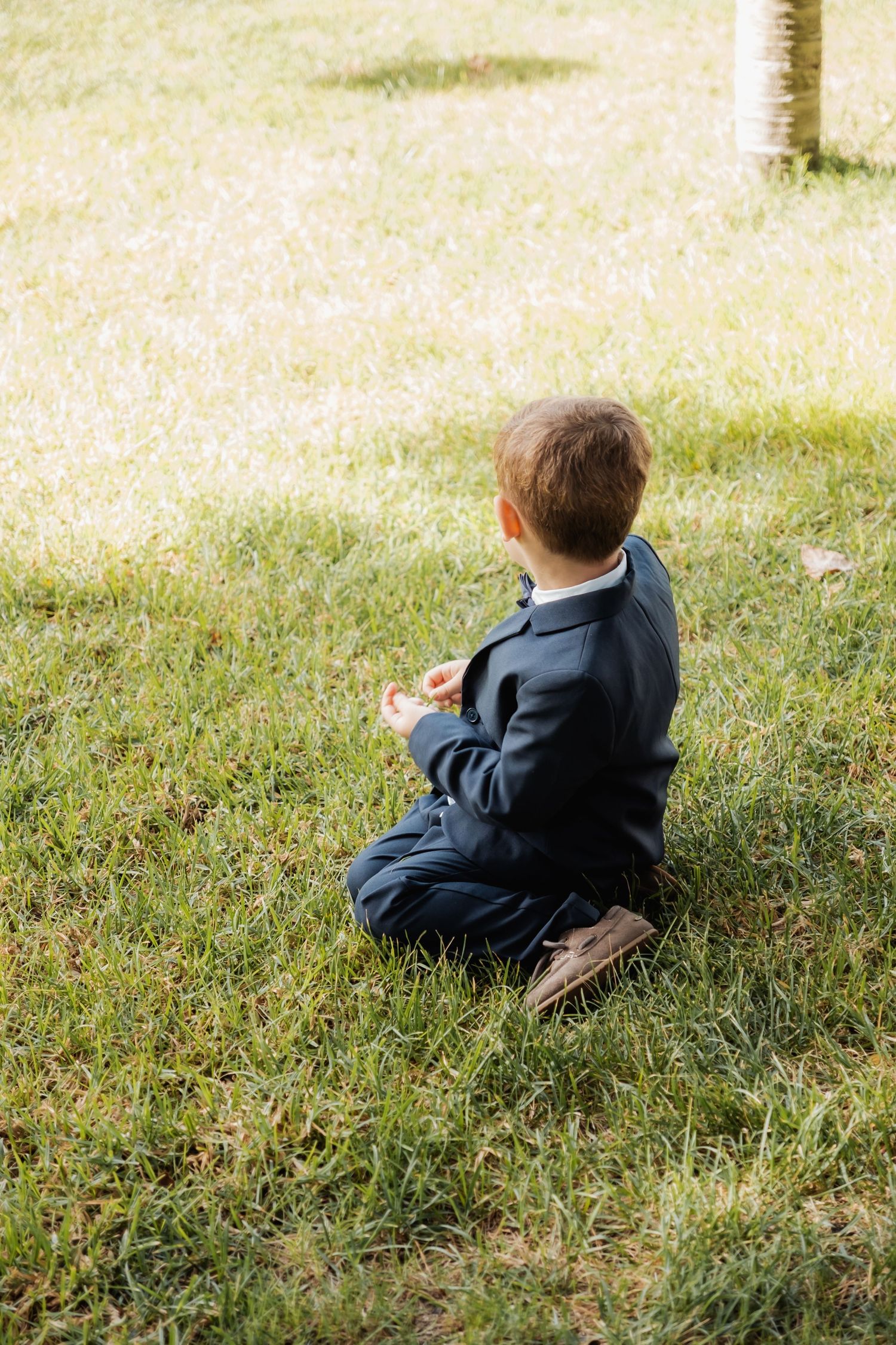 Young boy in navy suit sits cross-legged on green grass at outdoor wedding.