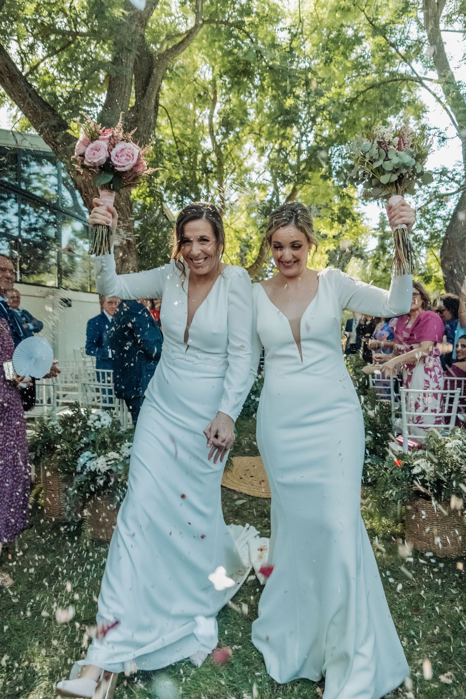Two joyful brides in white dresses raising bouquets as confetti falls during outdoor wedding exit.