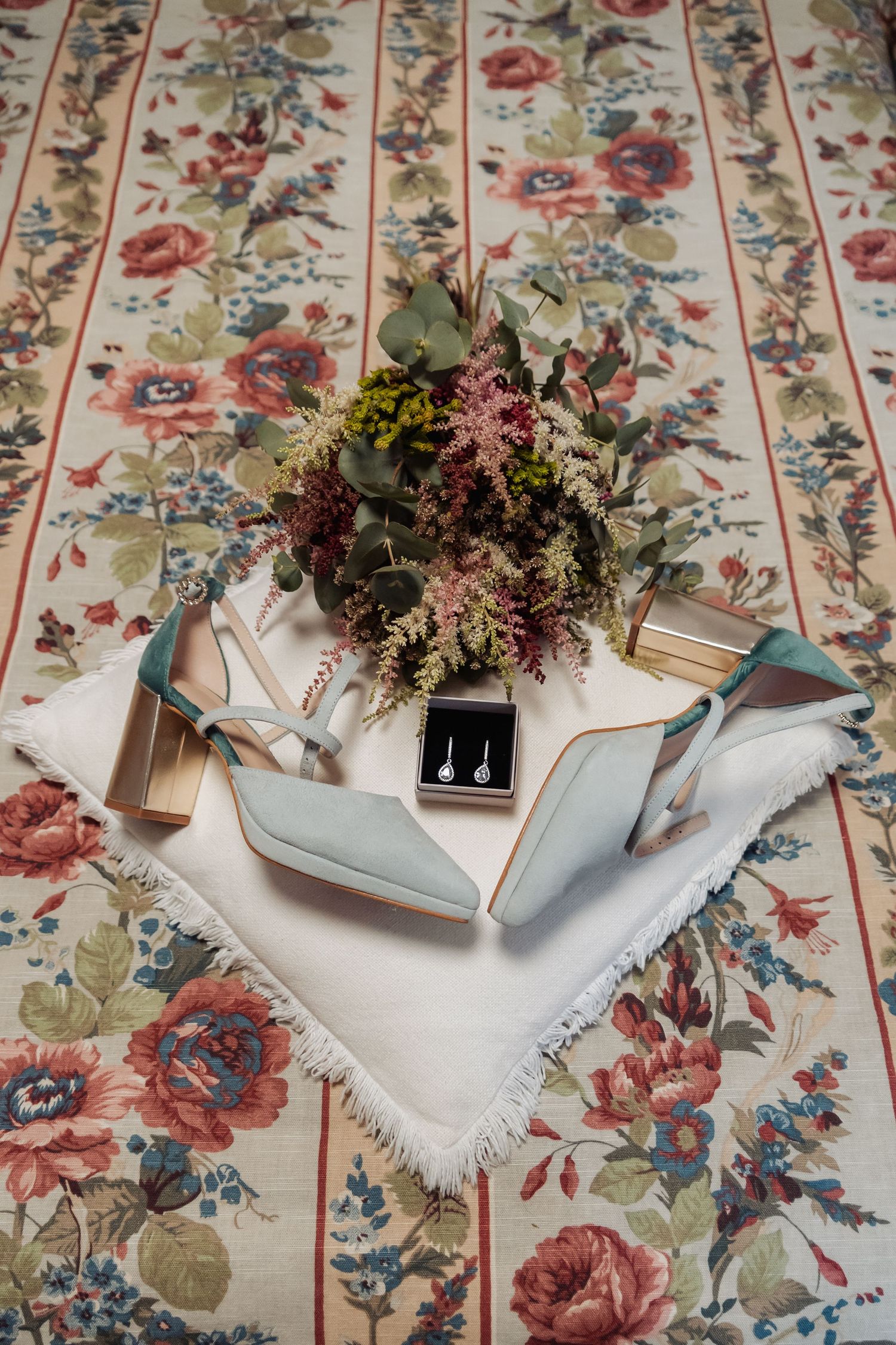 Wedding flat lay with blue heels, earrings, and floral bouquet on vintage floral tablecloth.