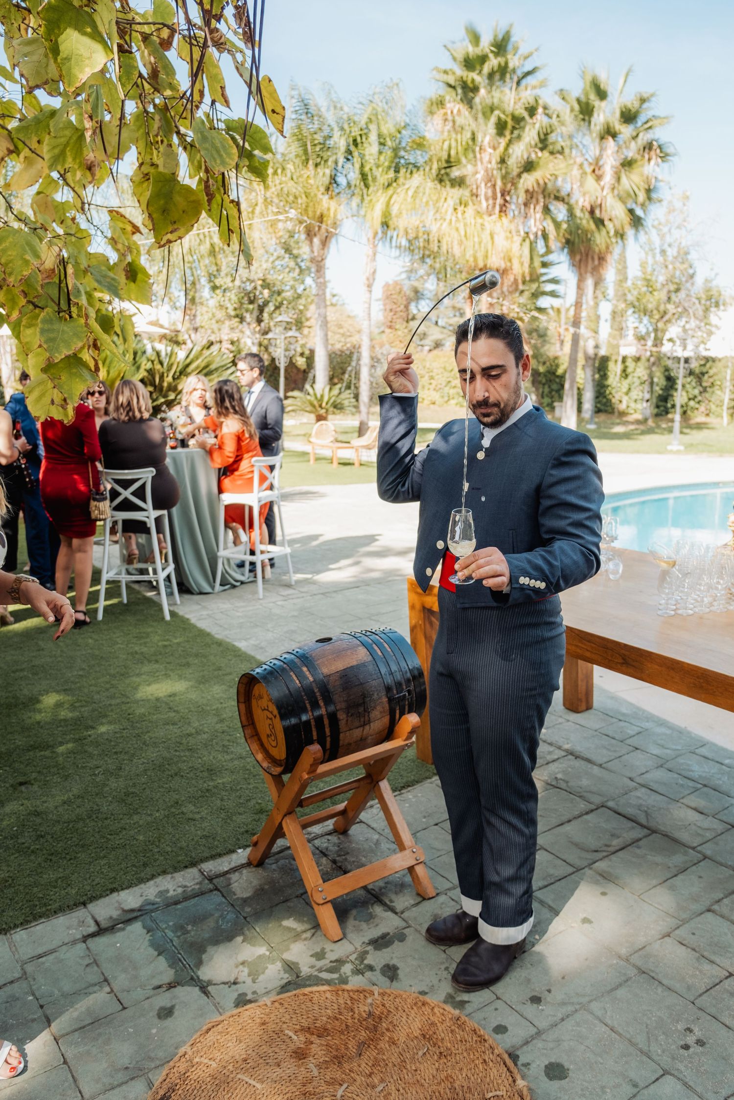 Man in suit pouring wine from a barrel at an outdoor wedding reception by a pool.
