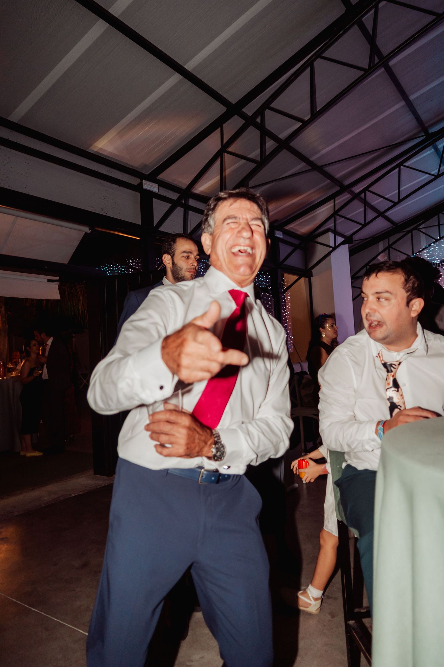 Energetic man in white shirt and red tie dancing and pointing at guests during a wedding reception.