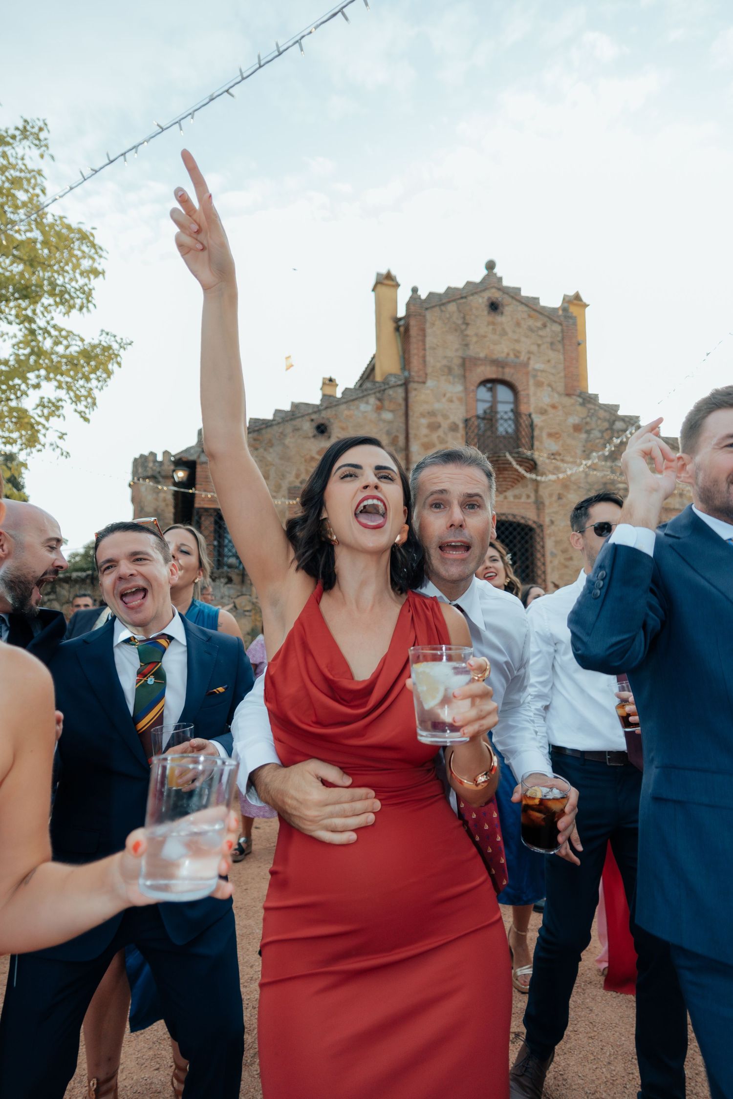 Woman in red dress celebrating with raised arm among wedding guests outside historic stone venue.