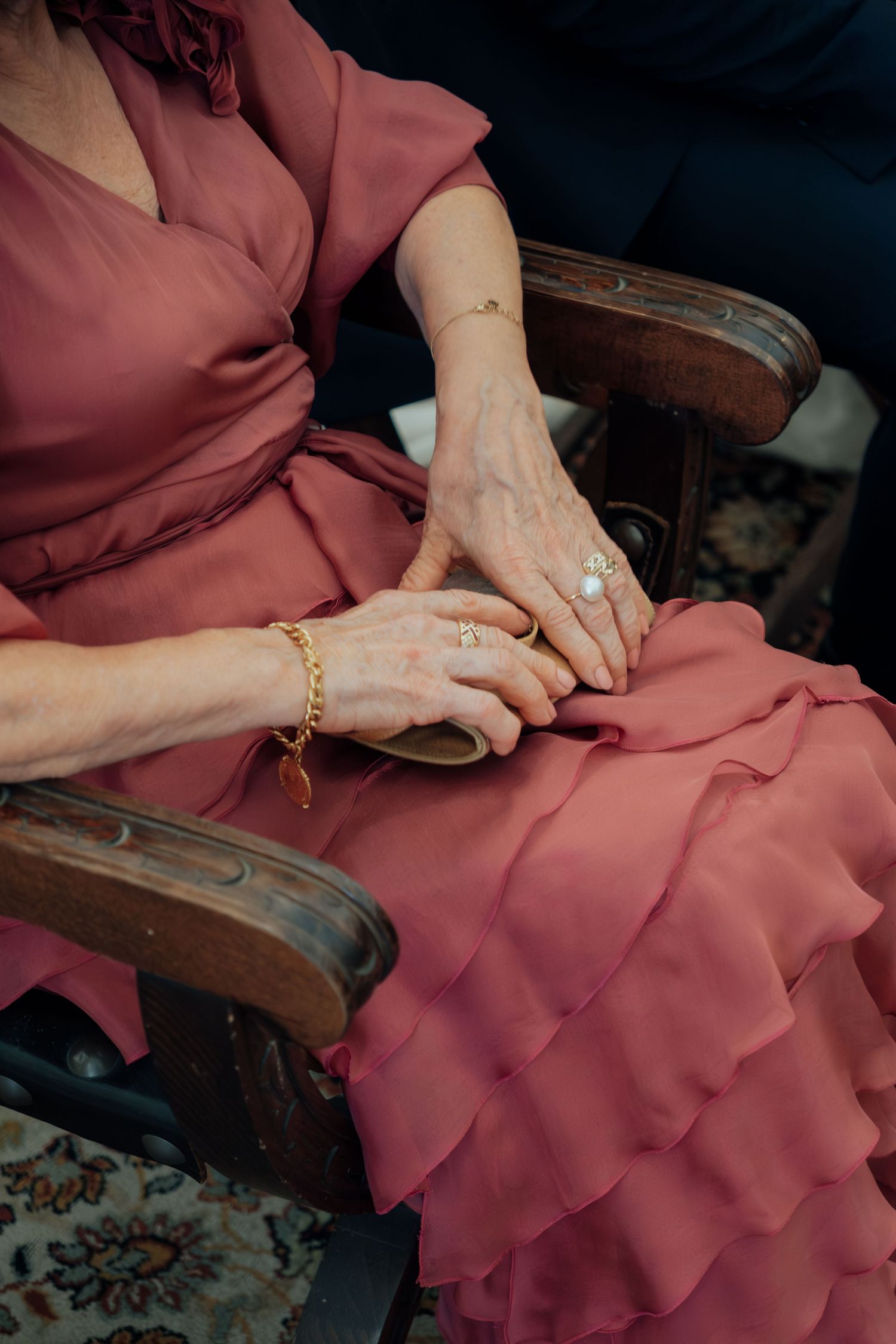 Close-up of an elegant woman in a pink ruffled dress wearing gold jewelry with clasped hands on her lap.