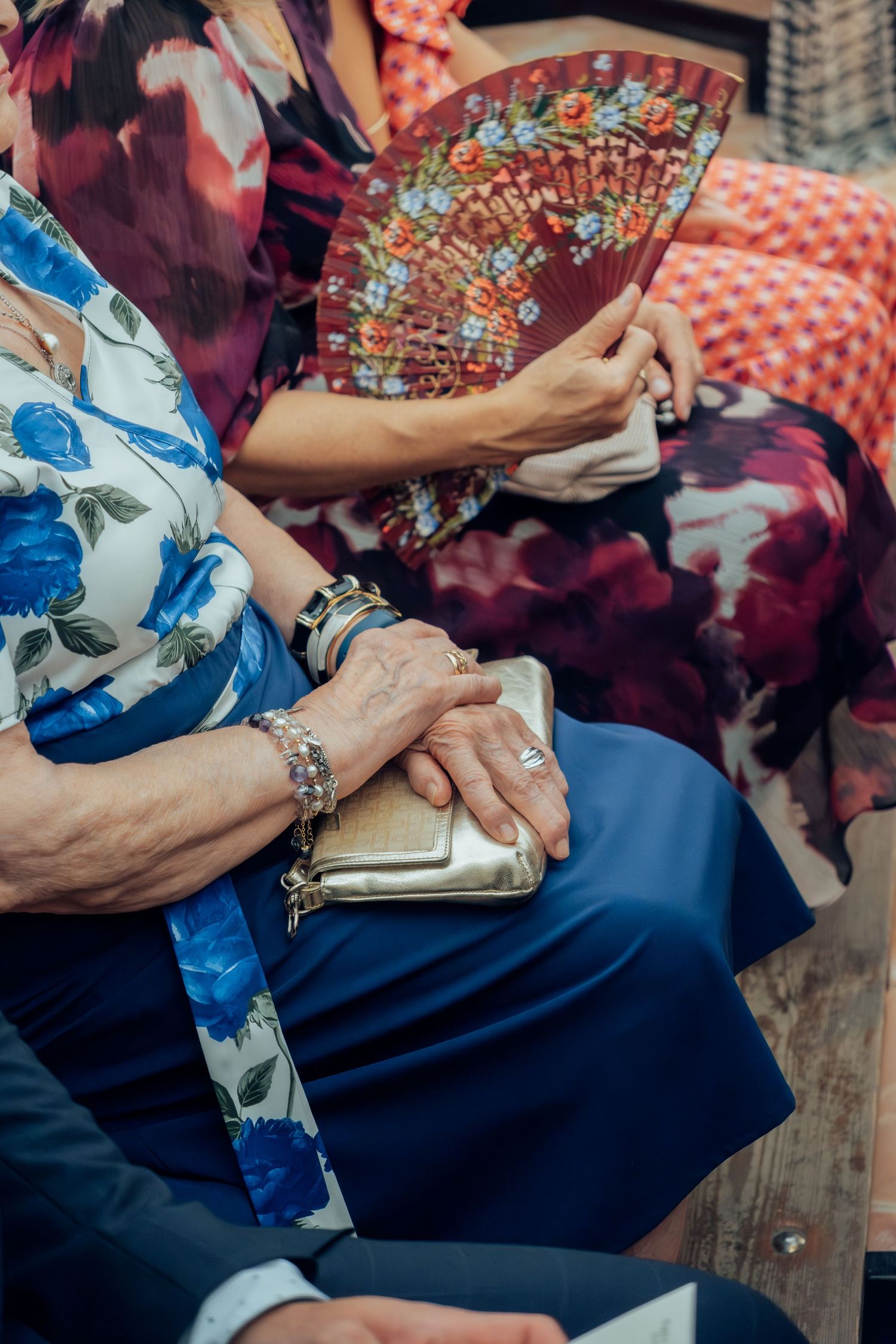 Elderly women seated at wedding holding decorative floral fan and golden clutch purse.