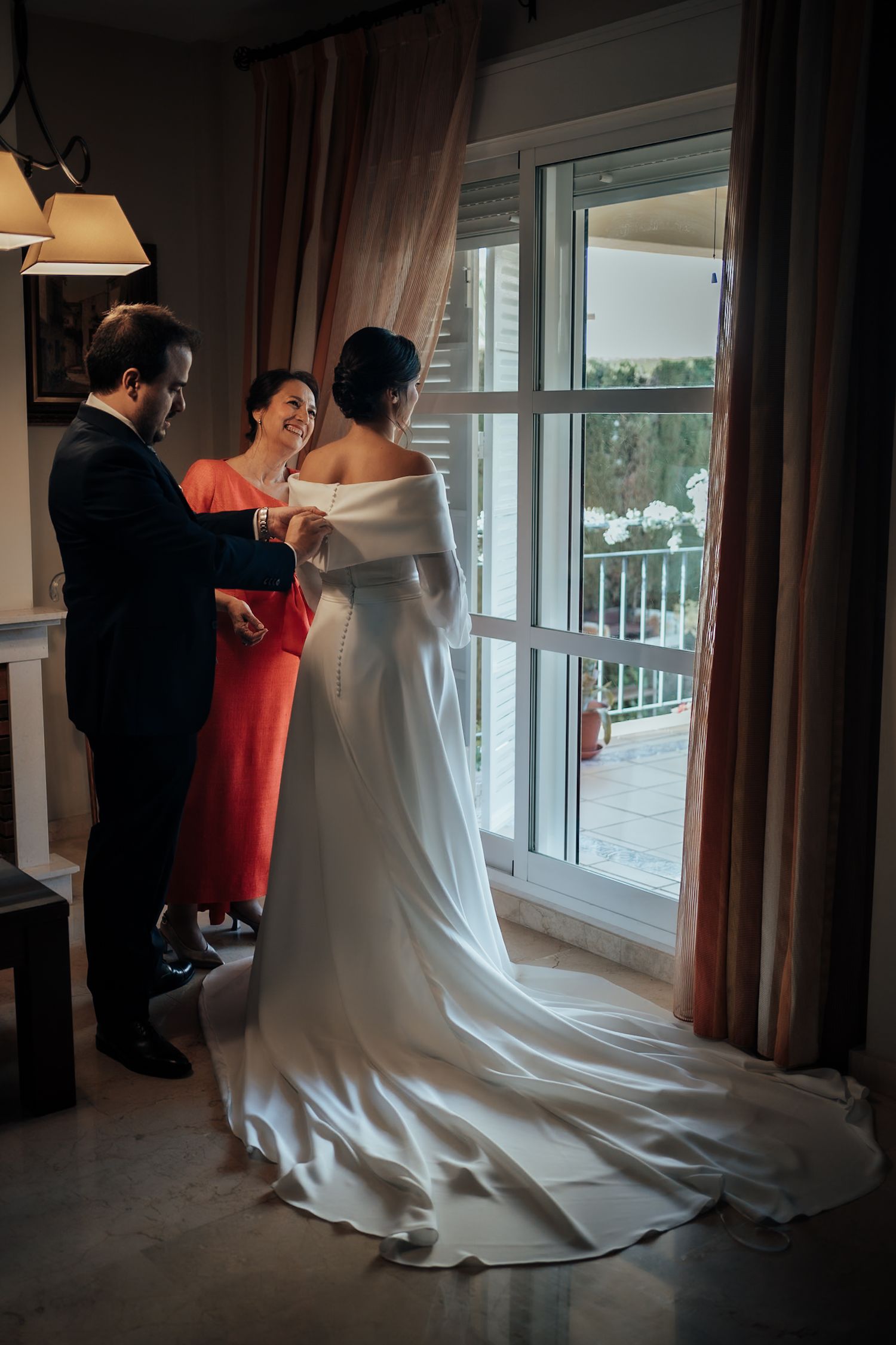 Bride in an off-shoulder white gown with a train being helped by family near a balcony window.