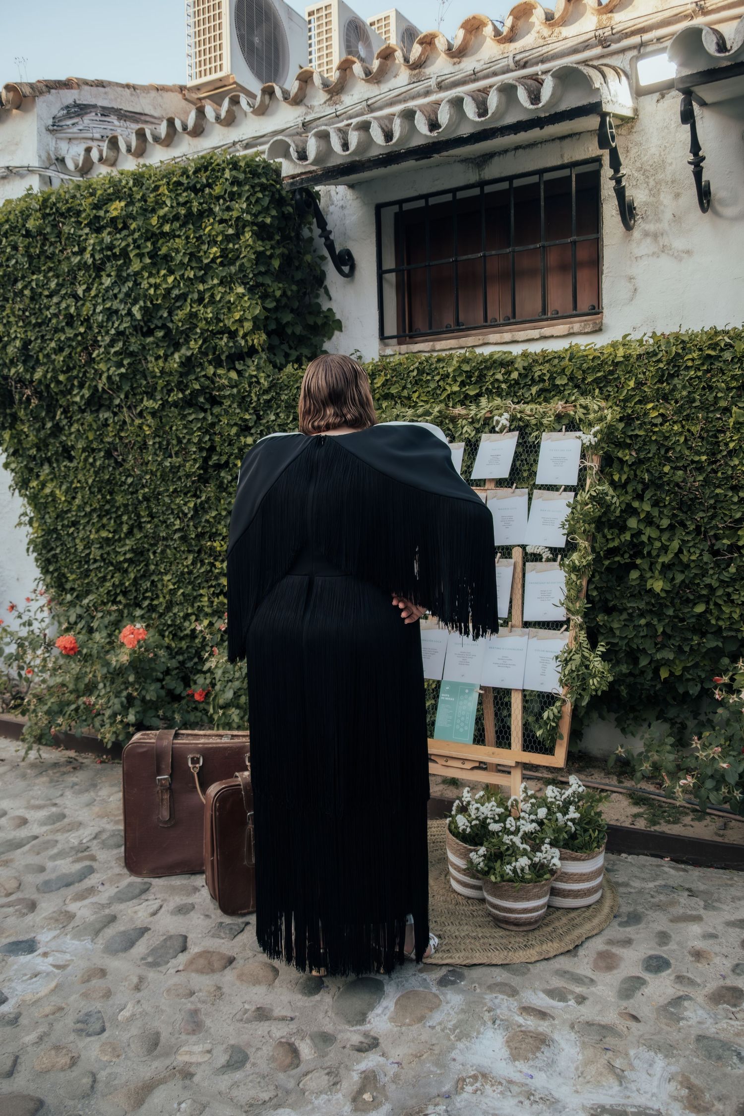 Woman in black fringed dress with vintage suitcase reviewing seating chart at ivy-covered venue entrance.