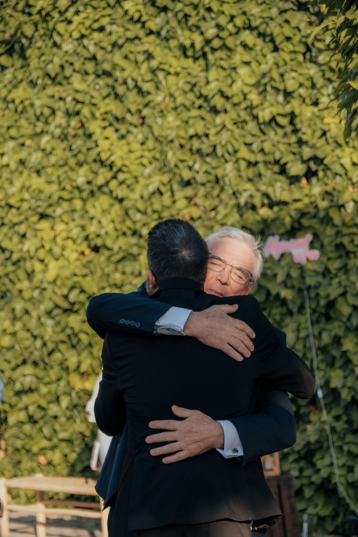Two men share an emotional embrace in front of lush green ivy wall at wedding celebration.