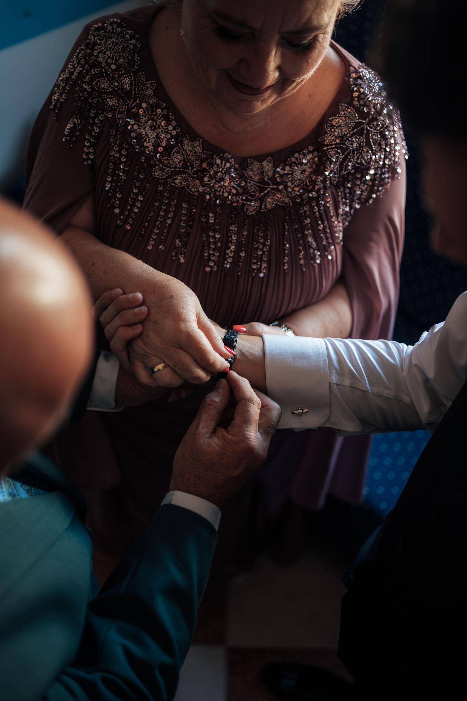 A smiling woman in a sequined dress helps fasten a cufflink on a groom's wrist.