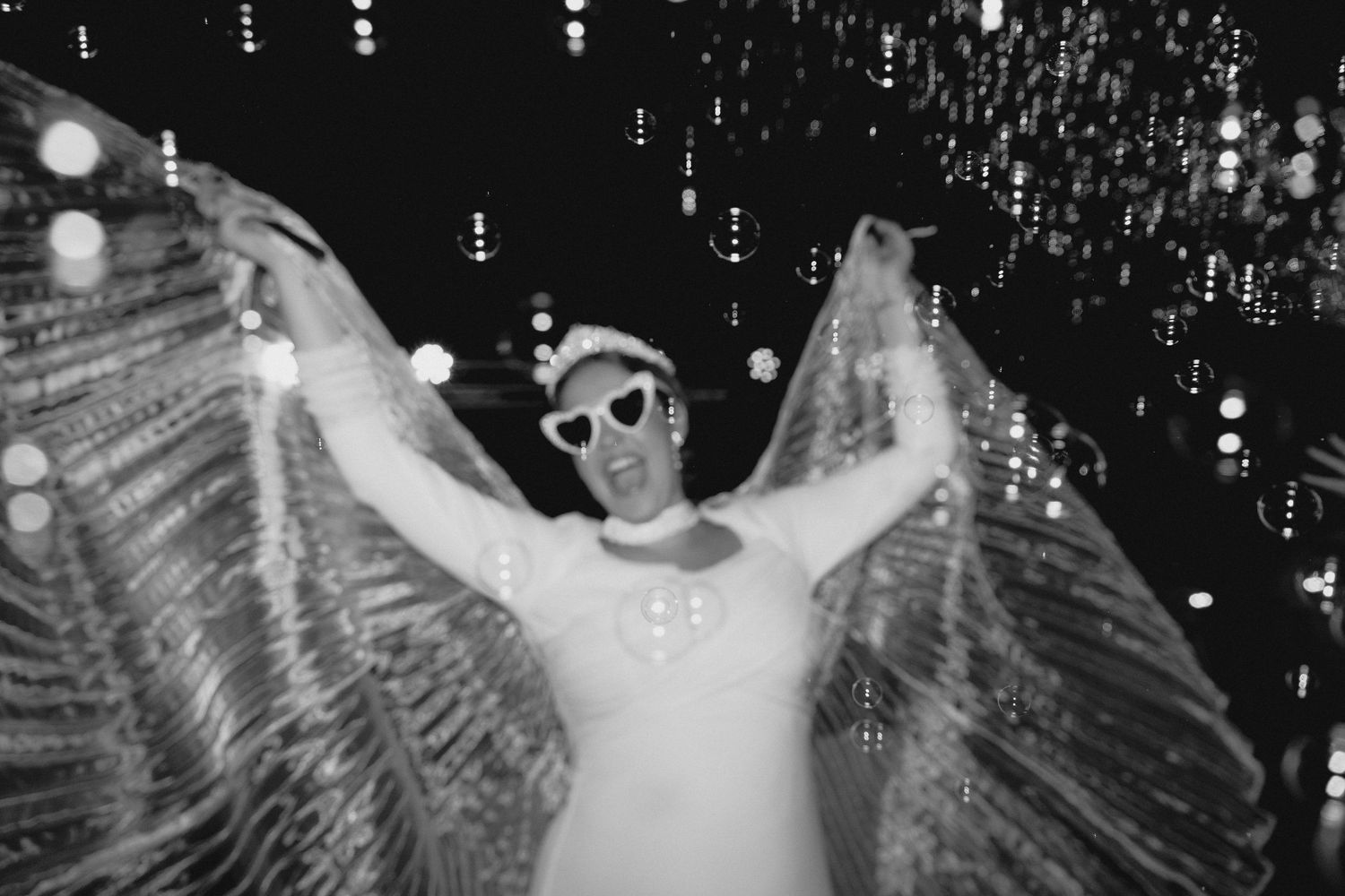 Black and white photo of a bride in white with sunglasses spreading glittery wings at night.