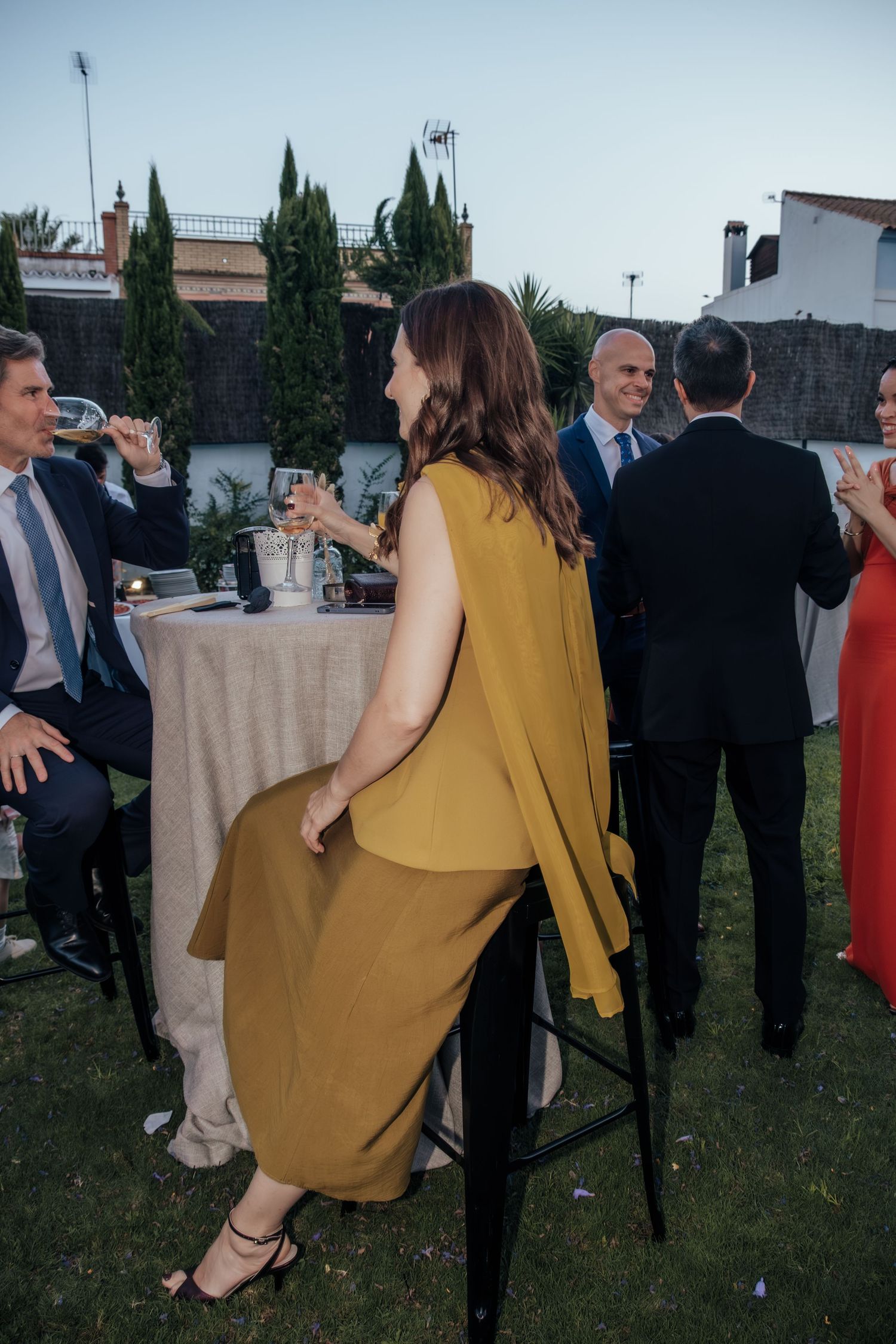 Woman in mustard yellow dress sitting at outdoor wedding cocktail hour event.