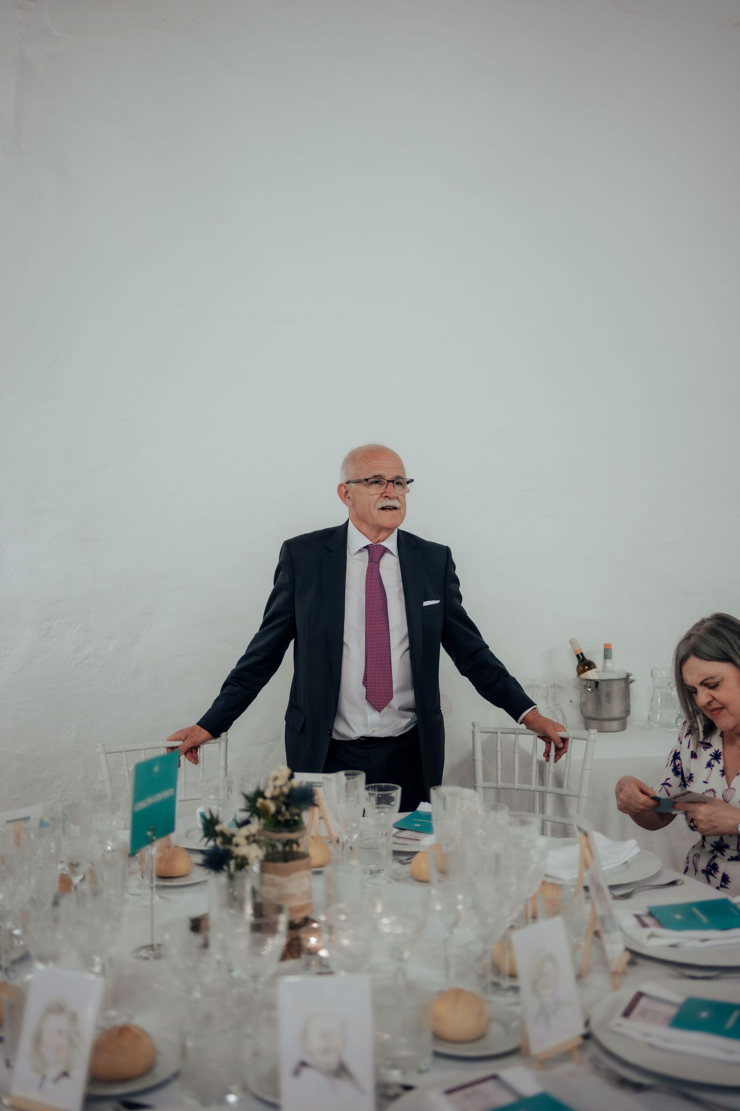 Elderly man in dark suit and pink tie standing at a elegantly set wedding reception table giving a speech.