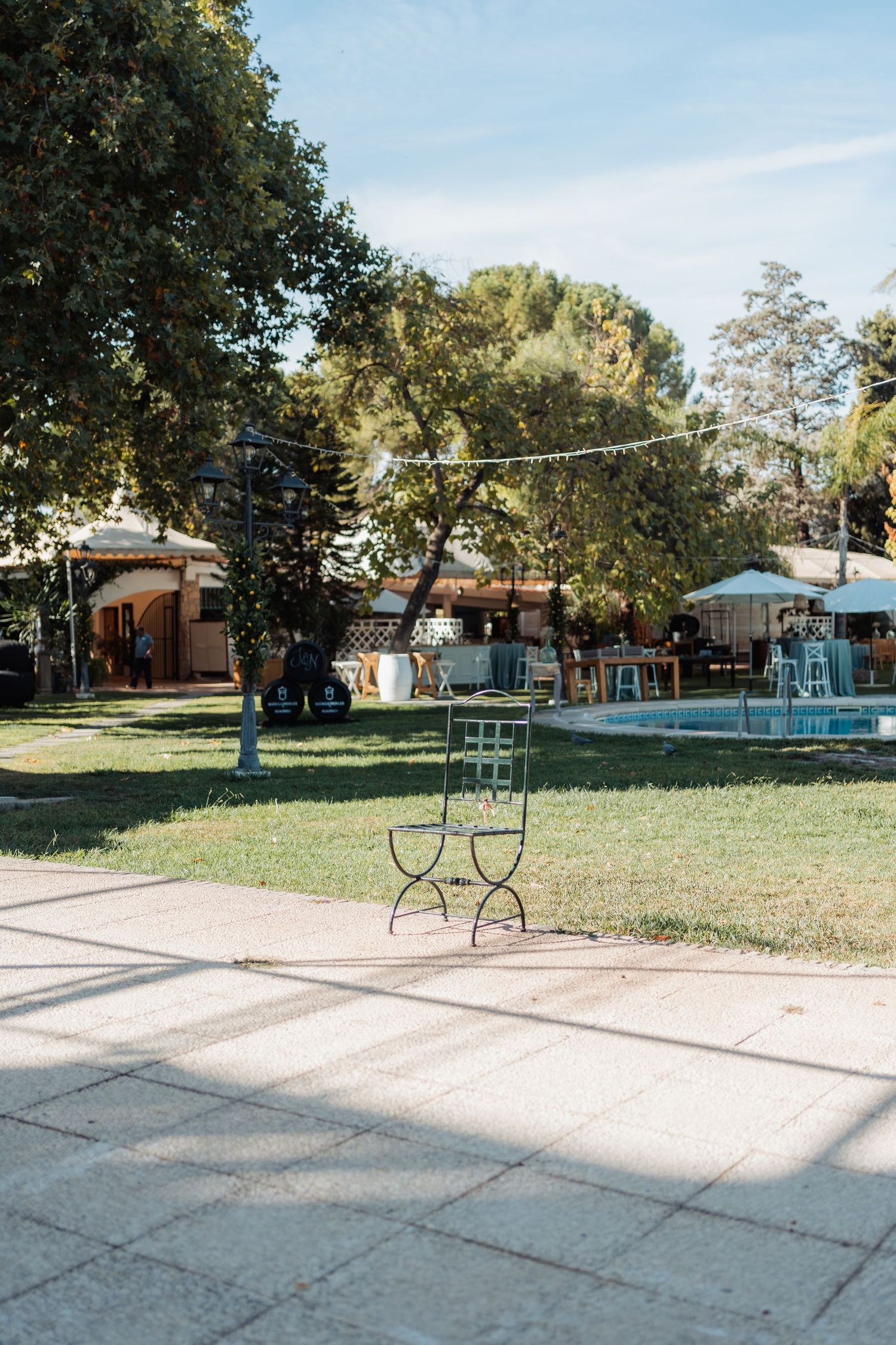 Sunlit garden wedding venue with green lawn, trees, string lights, and decorative metal chair.