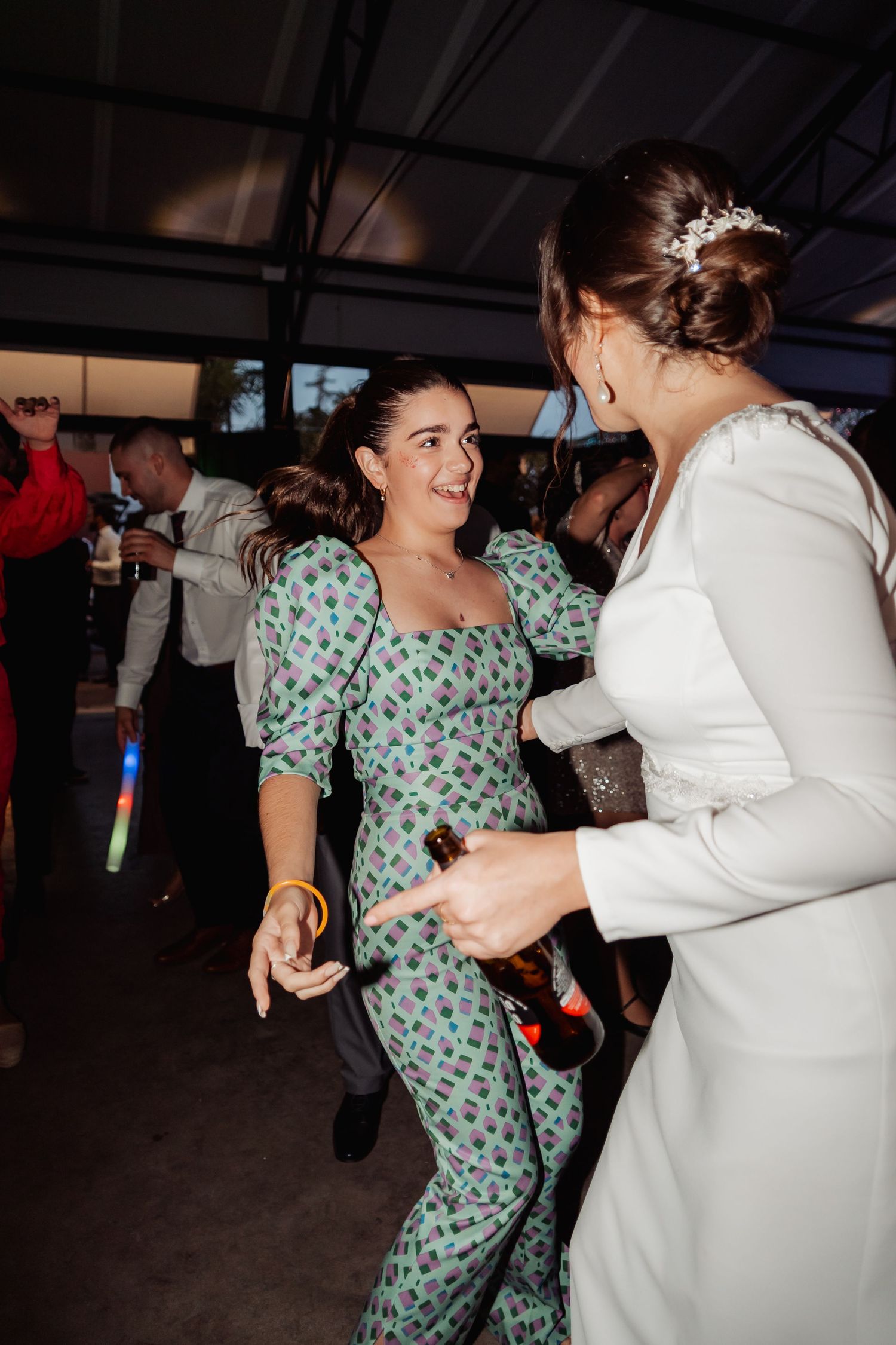 Guest in green patterned jumpsuit dancing joyfully with bride at a lively wedding reception.