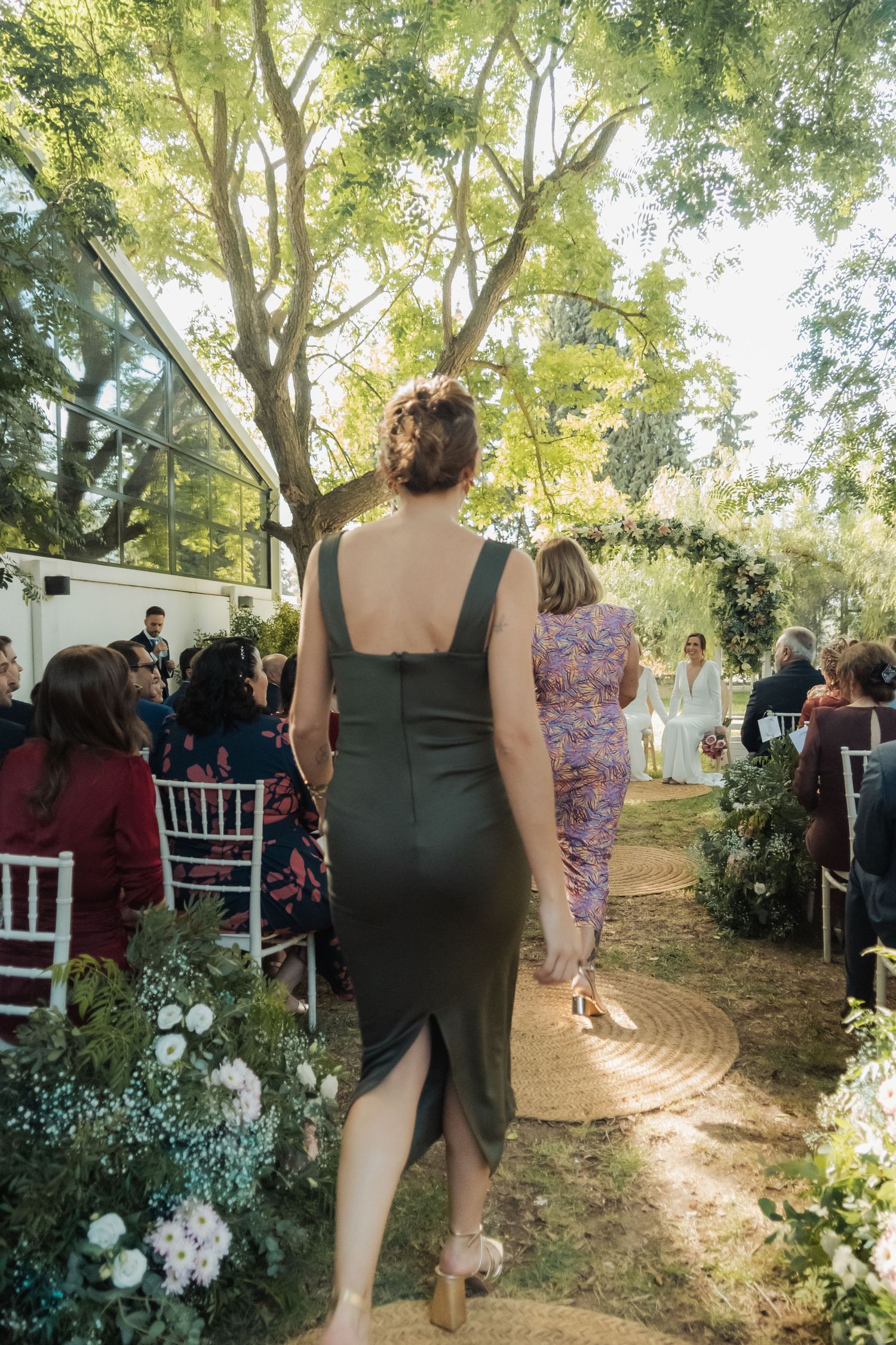 Woman in dark dress walks down sunlit outdoor wedding aisle toward floral arch altar.