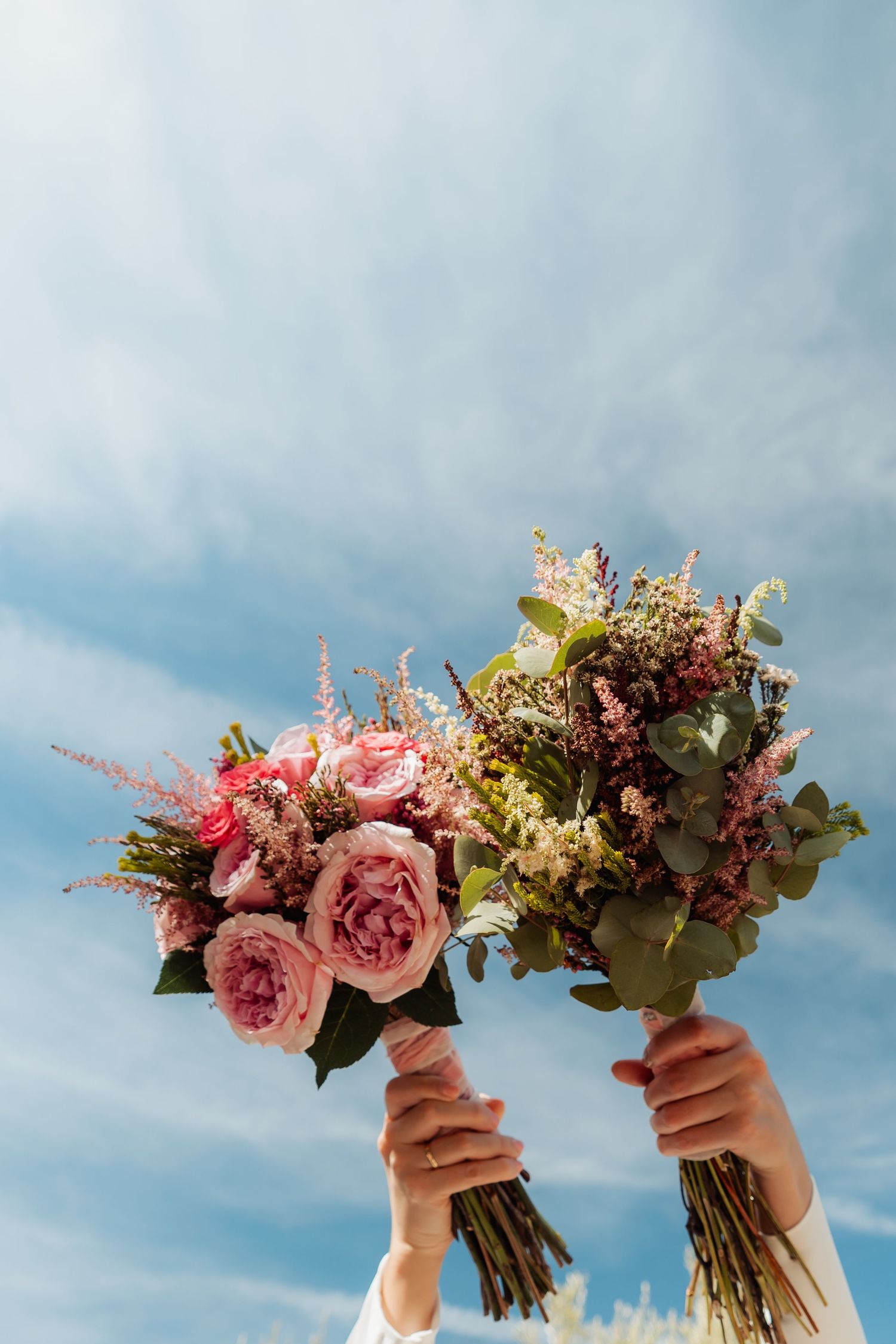 Two hands raise colorful wedding bouquets of pink roses and wildflowers against a blue sky.