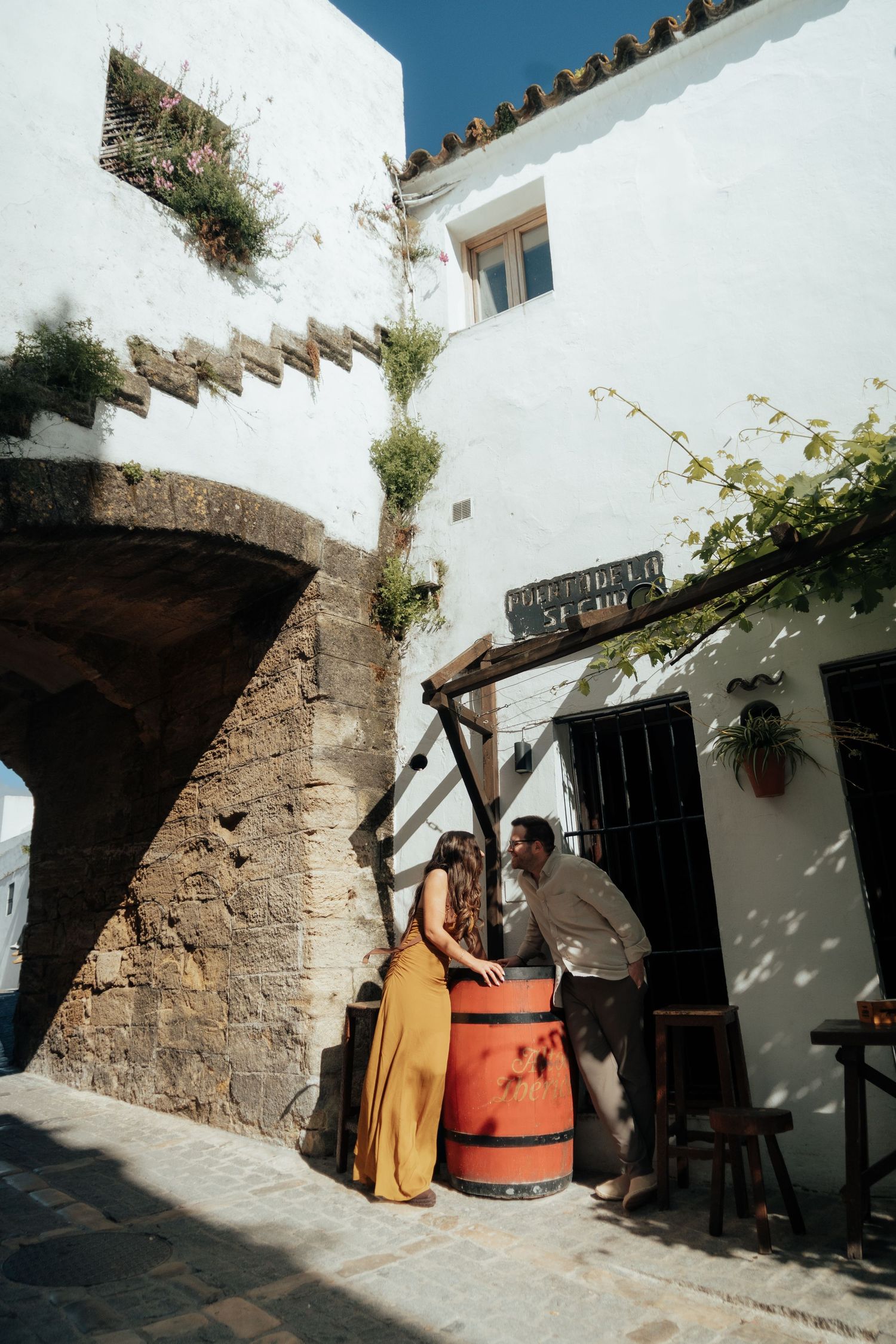 Couple in yellow dress and casual attire leaning on a red barrel in a charming white-walled Spanish alley.
