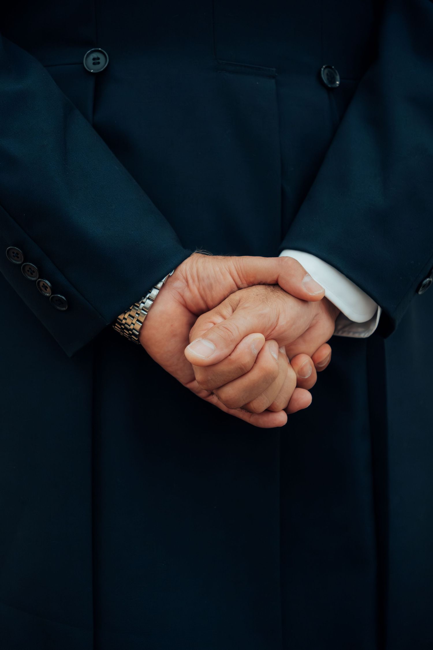 Close-up of man's clasped hands behind back wearing dark suit and gold watch at wedding.