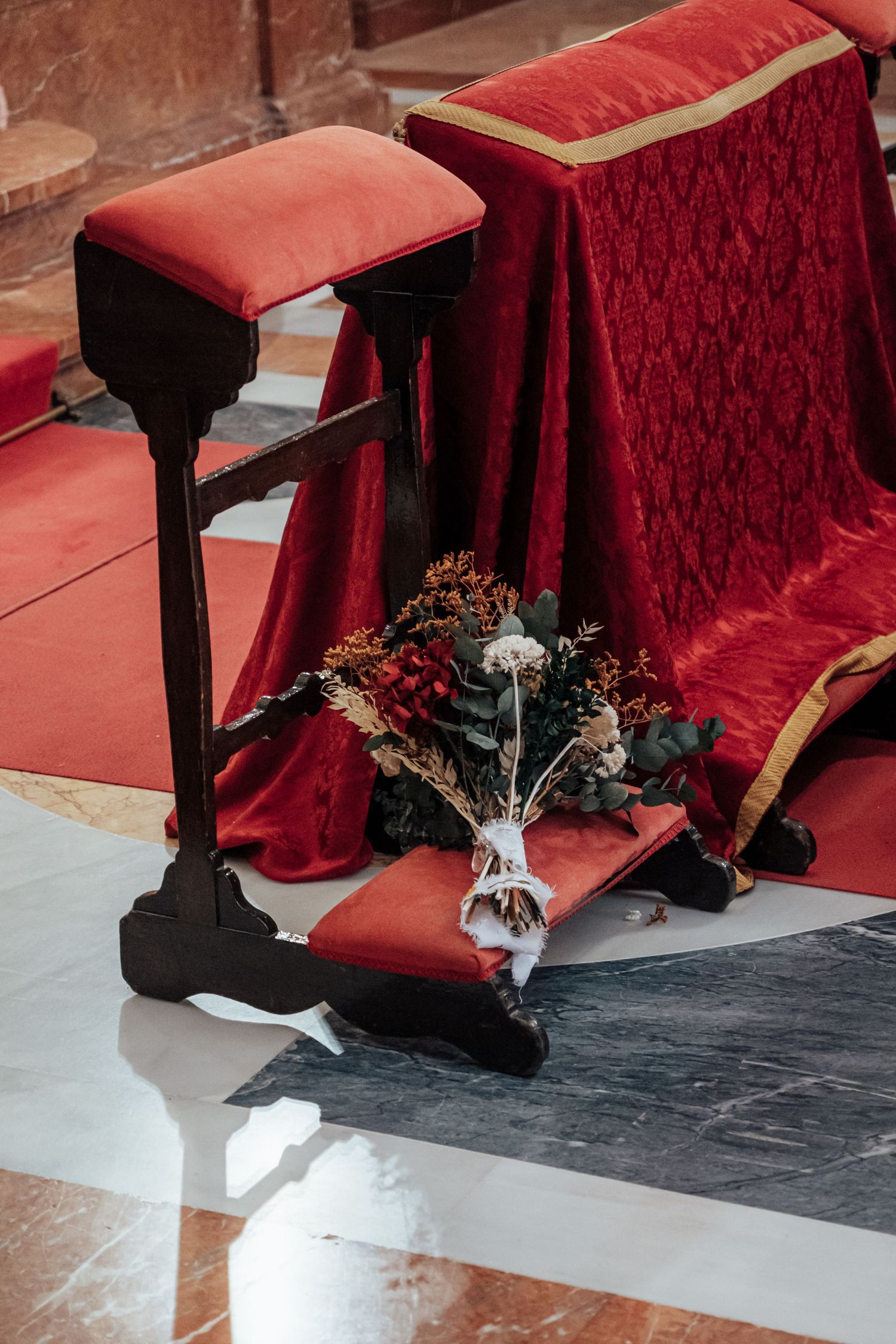 Wedding bouquet resting on red velvet kneeler beside wooden prie-dieu in church.