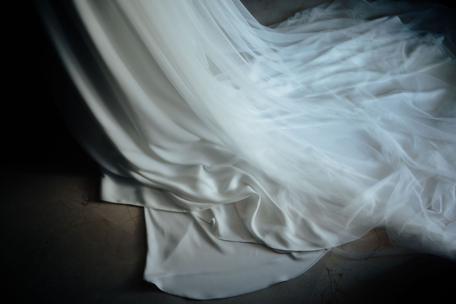 Flowing white wedding dress fabric with delicate tulle layers draped beautifully on the floor in soft dramatic lighting.