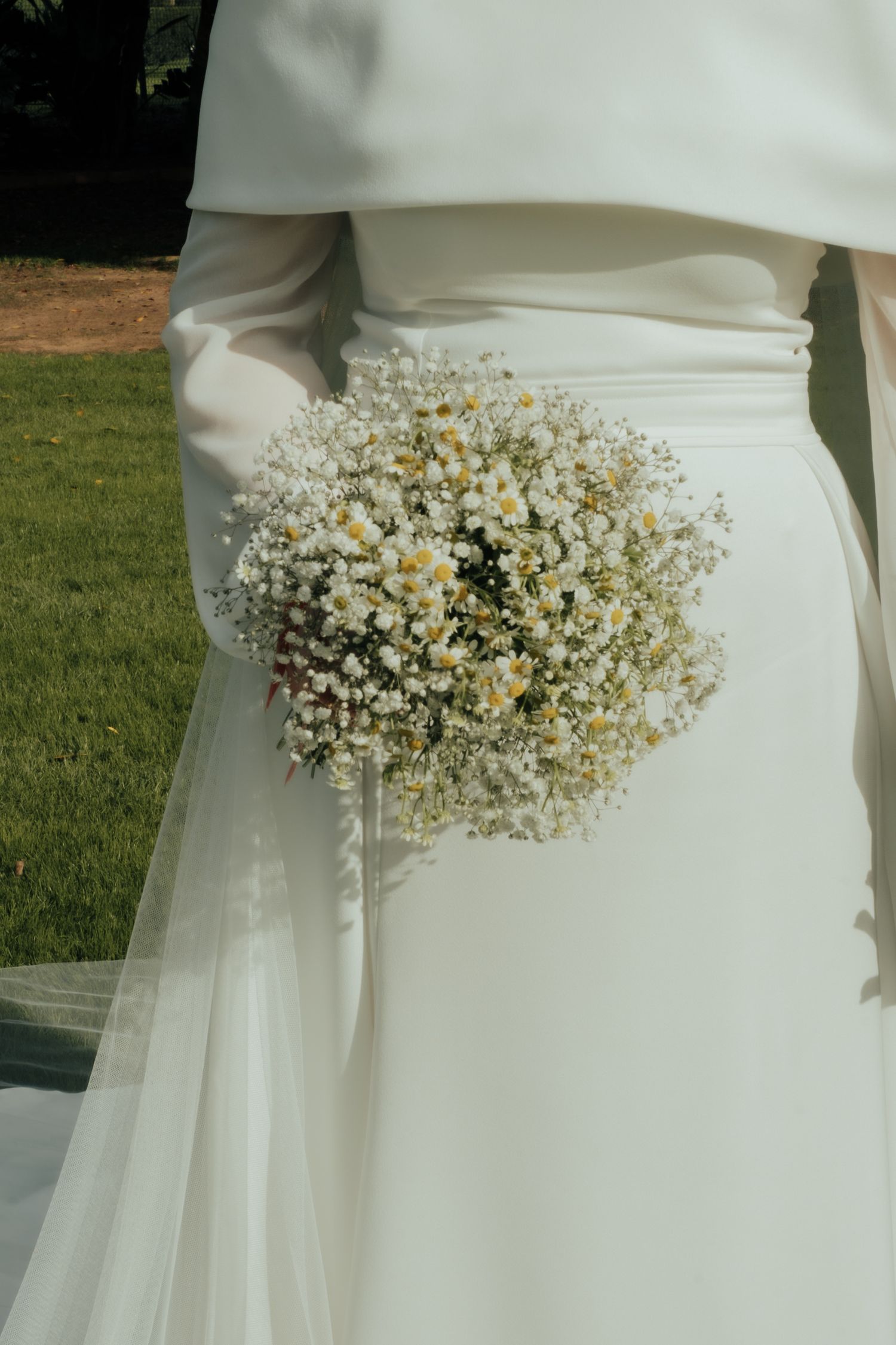 Close-up of bride holding delicate white daisy and baby's breath bouquet against flowing white wedding gown.