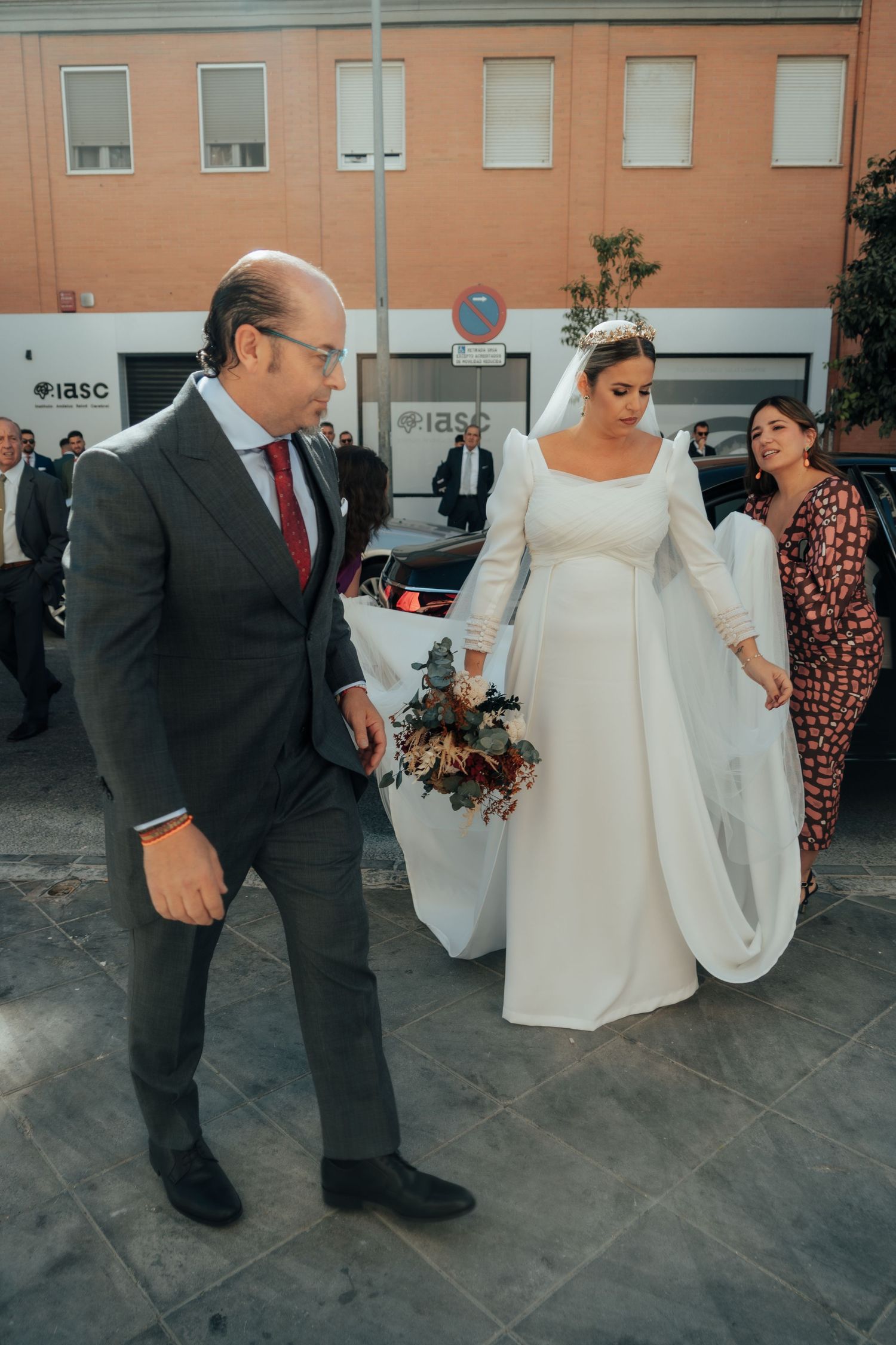 Bride in elegant white long-sleeve gown holding bouquet arriving at church with escort.