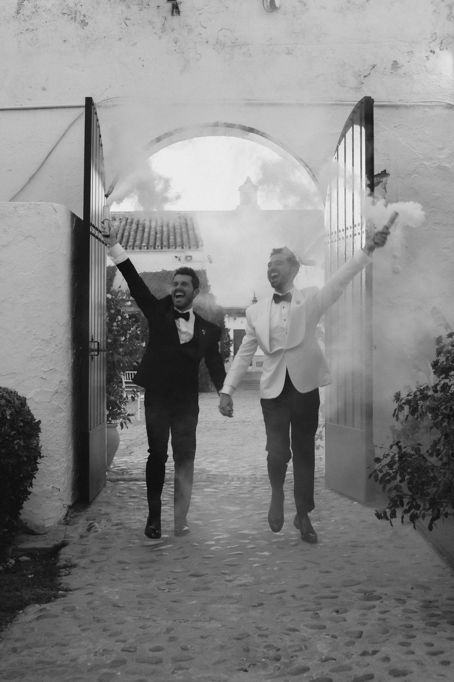 Black and white photo of two grooms in tuxedos celebrating with smoke flares through arched stone gateway.