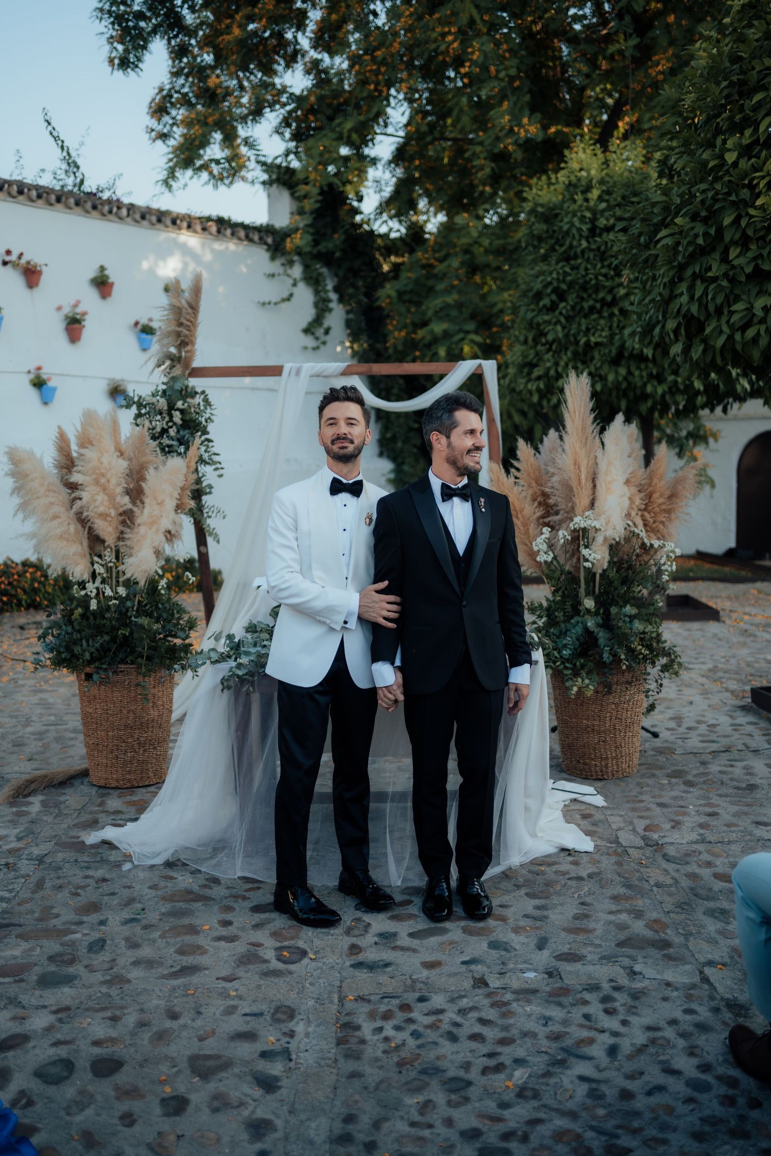 Two grooms in tuxedos pose before floral arch with pampas grass at outdoor wedding ceremony.