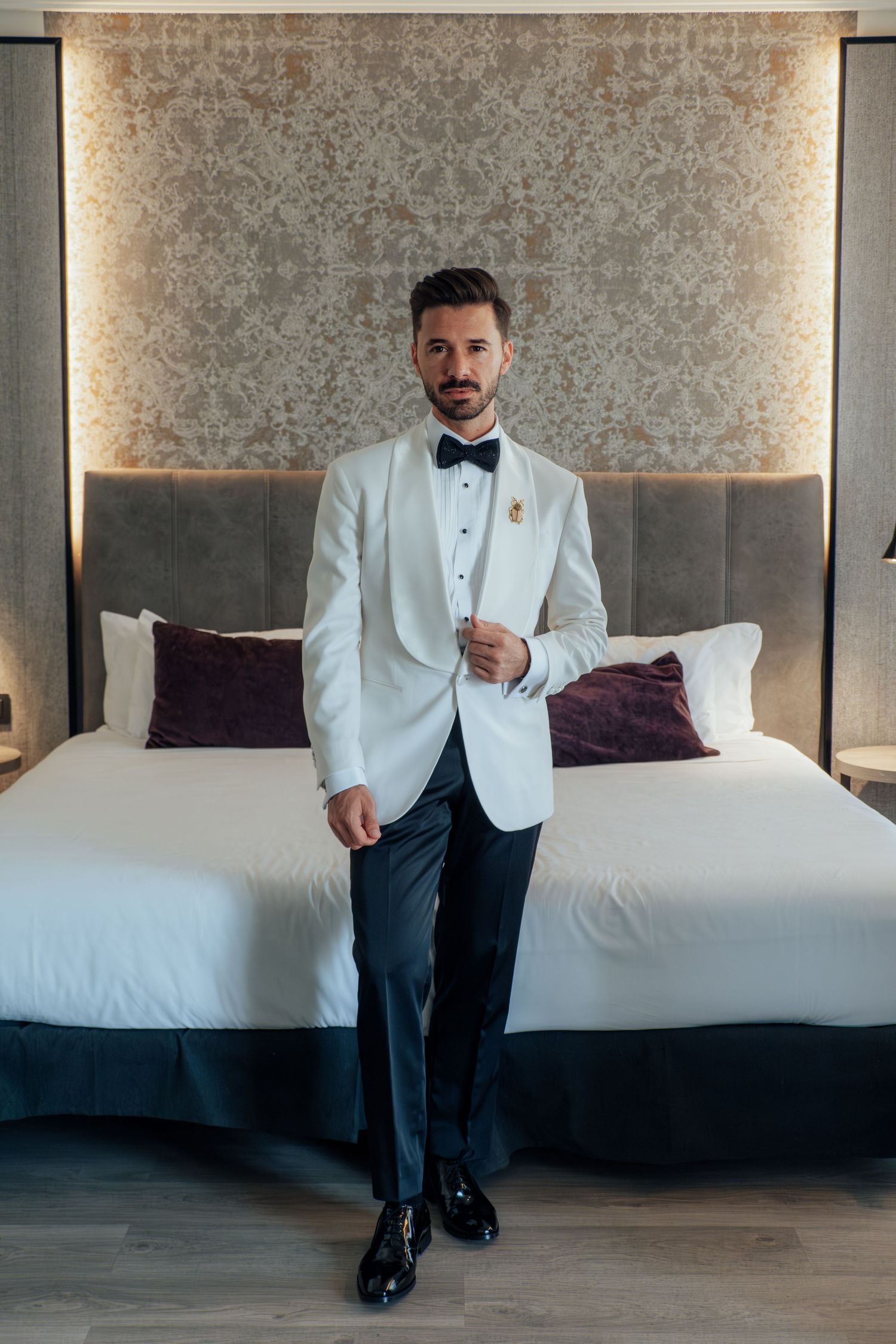 Dapper groom in white tuxedo with boutonniere stands confidently in a stylish hotel room.
