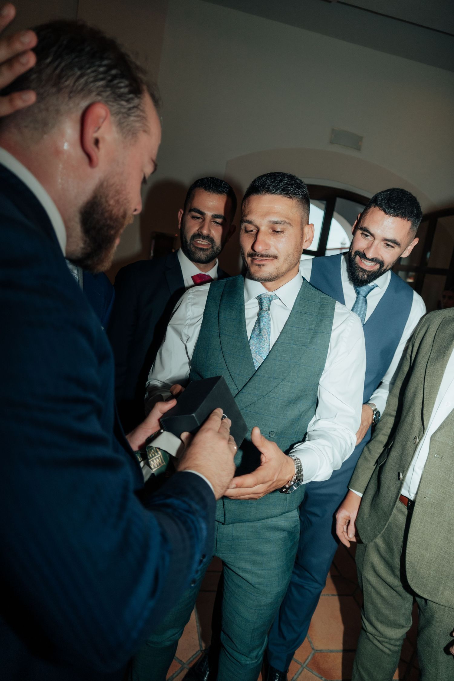 Smiling groomsmen in suits gather around man holding small gift box at wedding celebration.