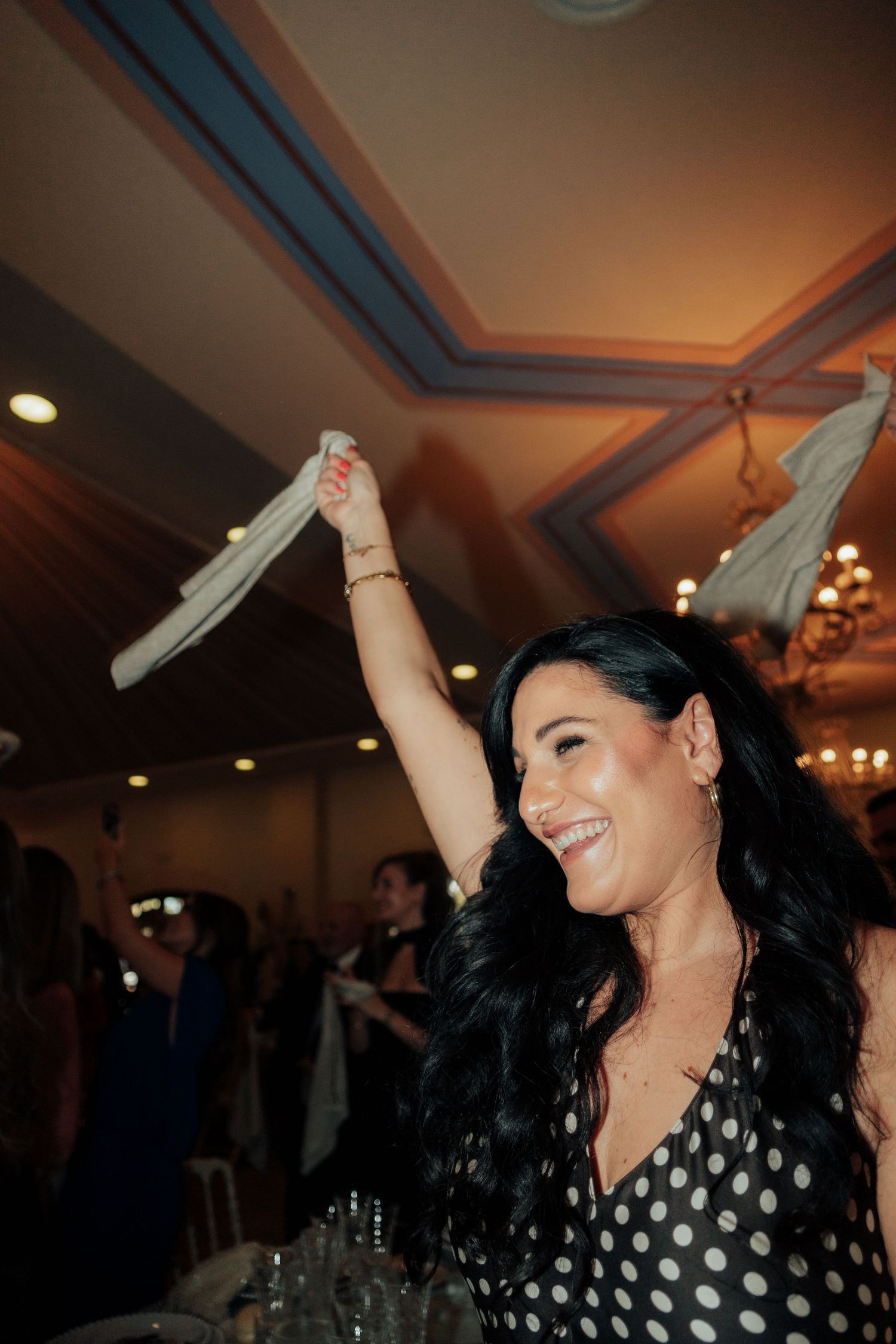 Joyful dark-haired woman in polka dot dress waving a napkin at a lively wedding reception.