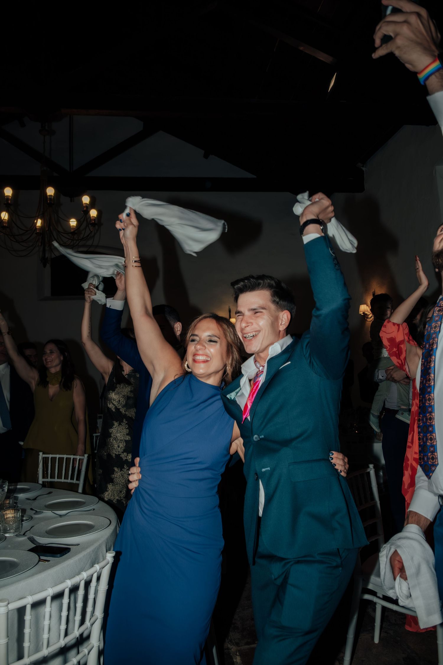 Joyful couple in blue dress and teal suit waving white napkins on wedding reception dance floor.