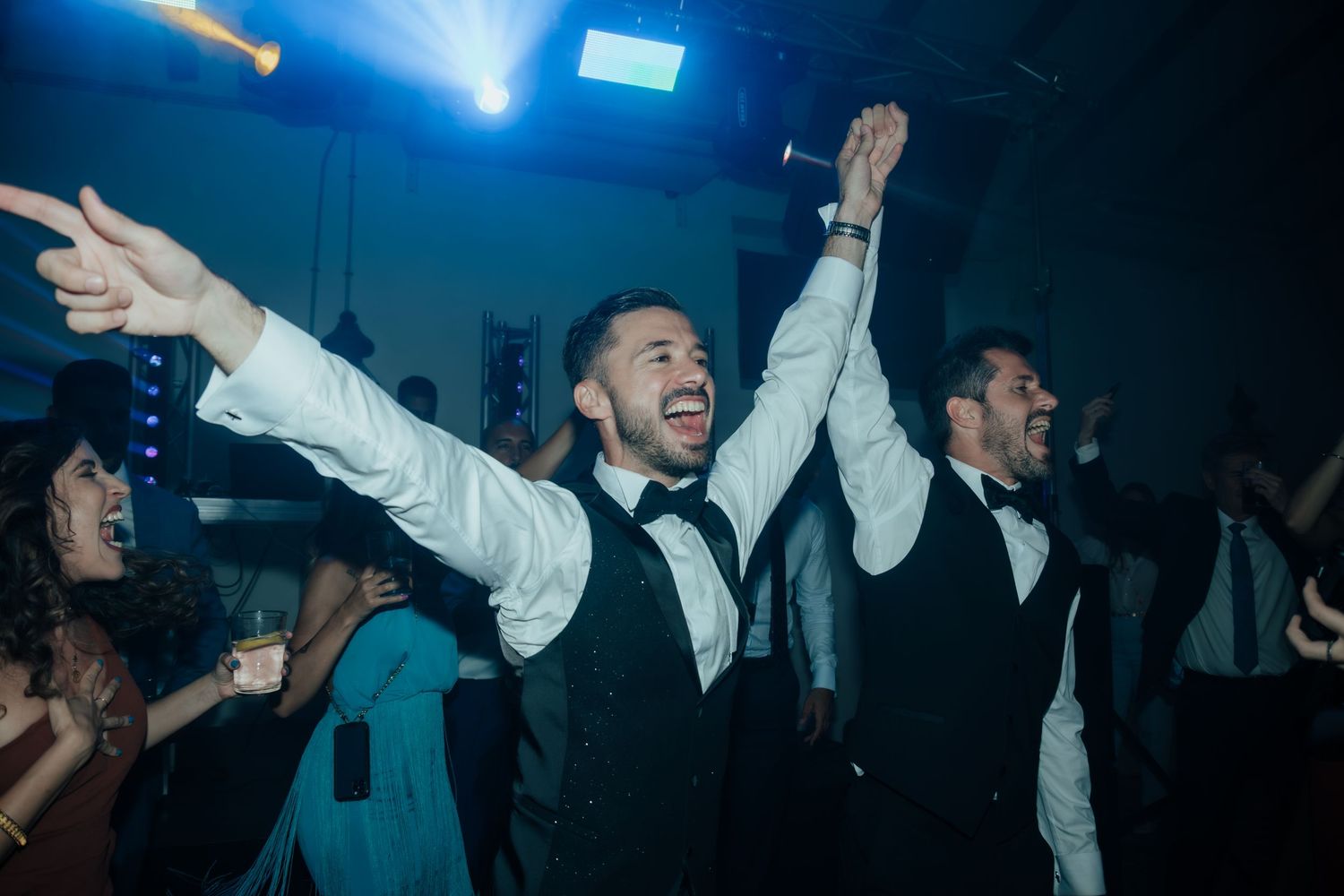 Two men in black vests and bow ties joyfully dancing with arms raised at a wedding reception party.