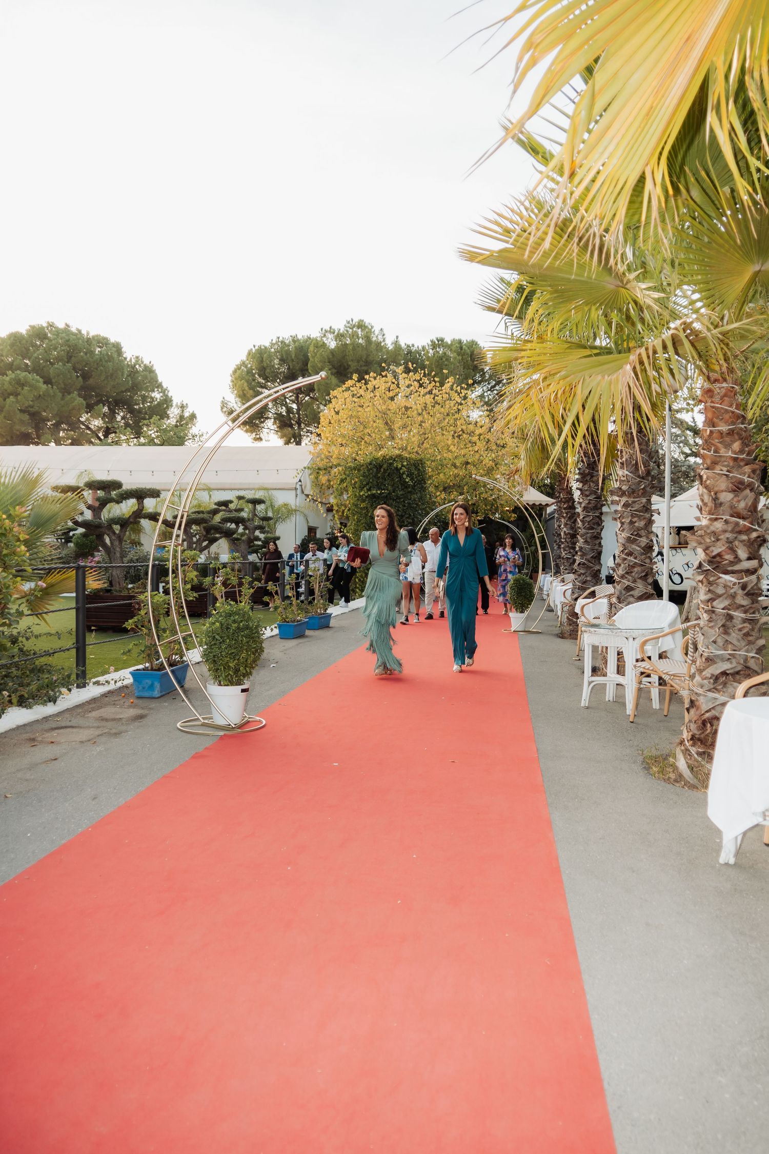 Guests walk along a red carpet lined with palm trees and circular floral arches at an outdoor event.