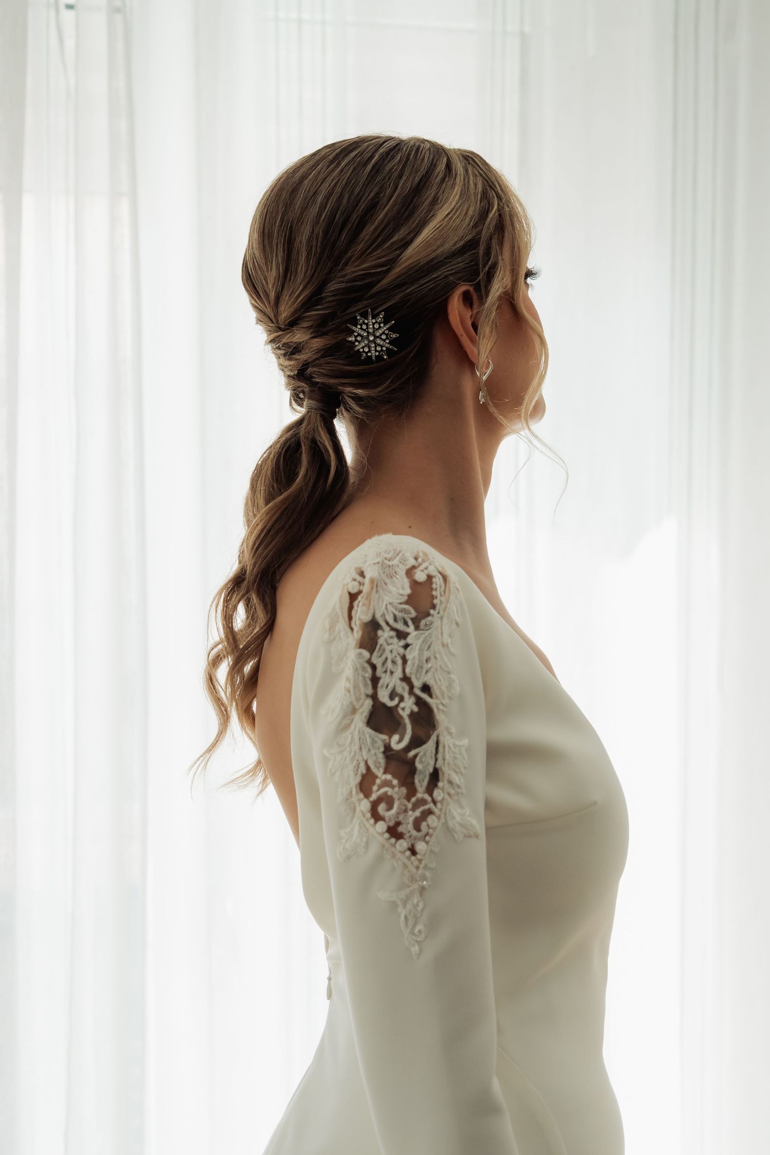 Side profile of bride with ponytail, crystal hair pin, and lace sleeve wedding dress.