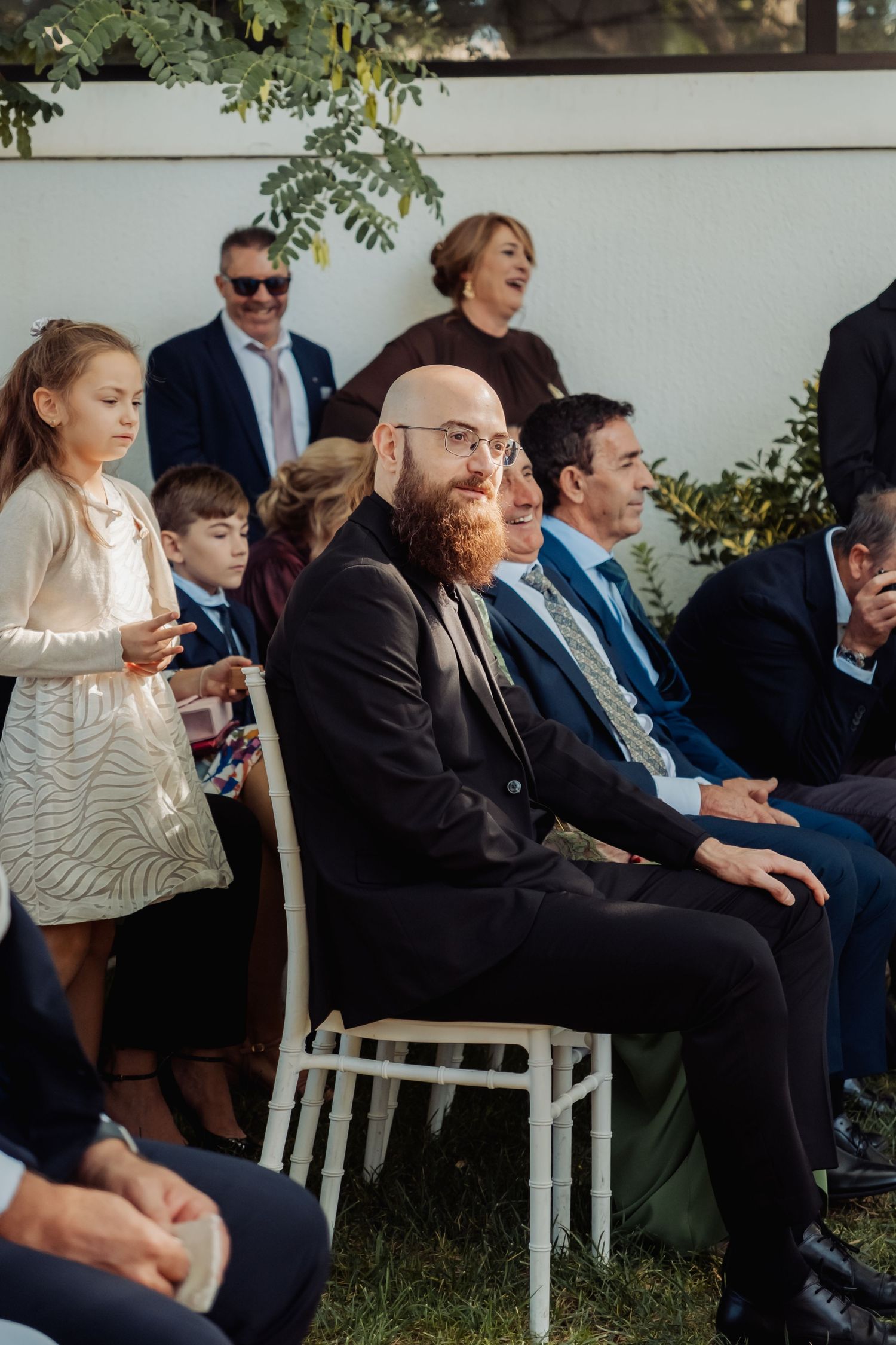 Bald bearded man in suit smiles while seated among wedding guests at outdoor ceremony.